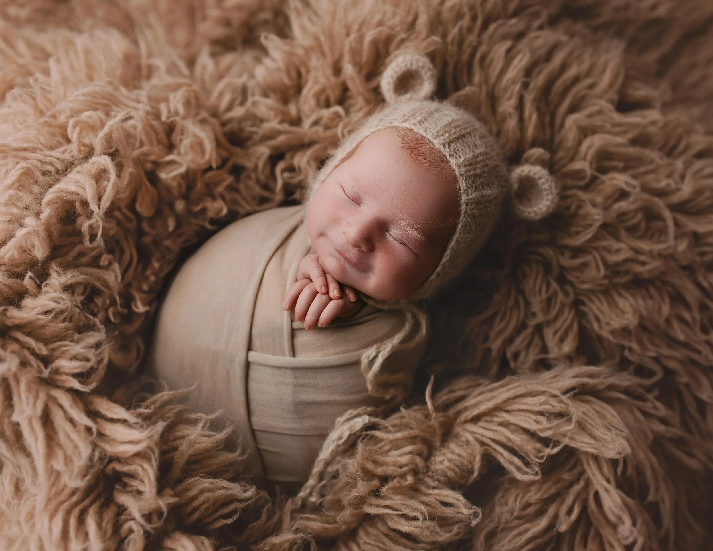 Smilende baby med nisselue hviler på en myk, brun, fluffy pute, pakket inn i beige klær.