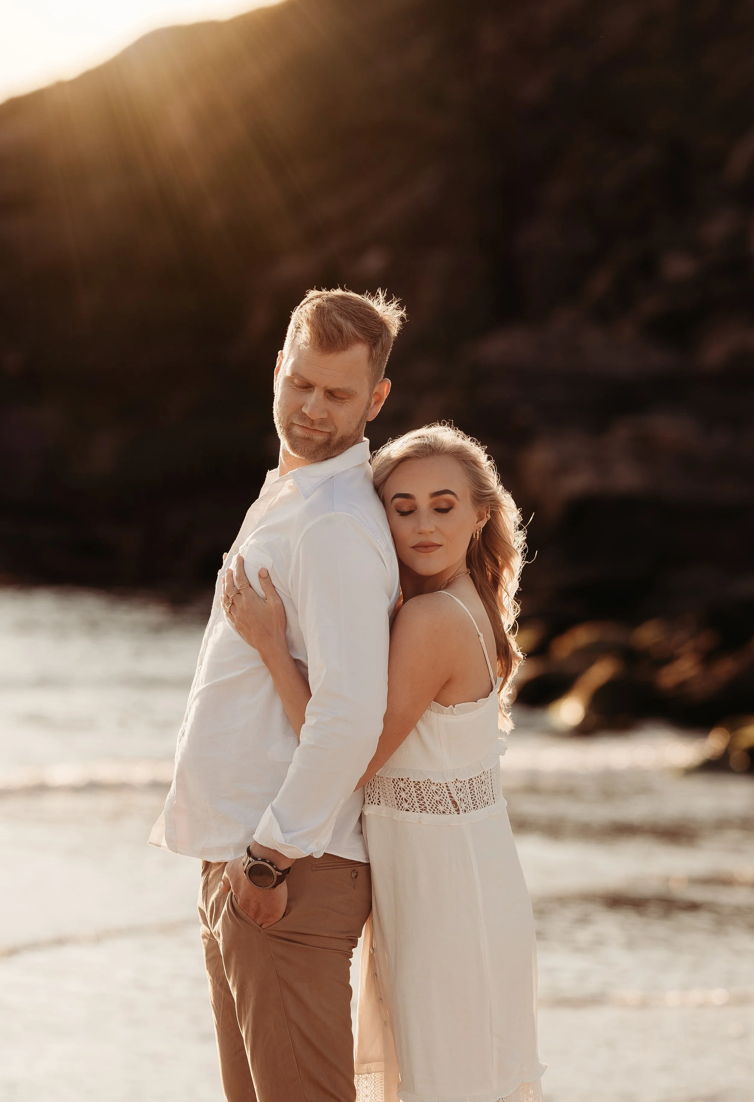 Et vakkert bilde av et par som står på stranden med hav og fjell i bakgrunnen, med solnedgang i bakgrunnen.