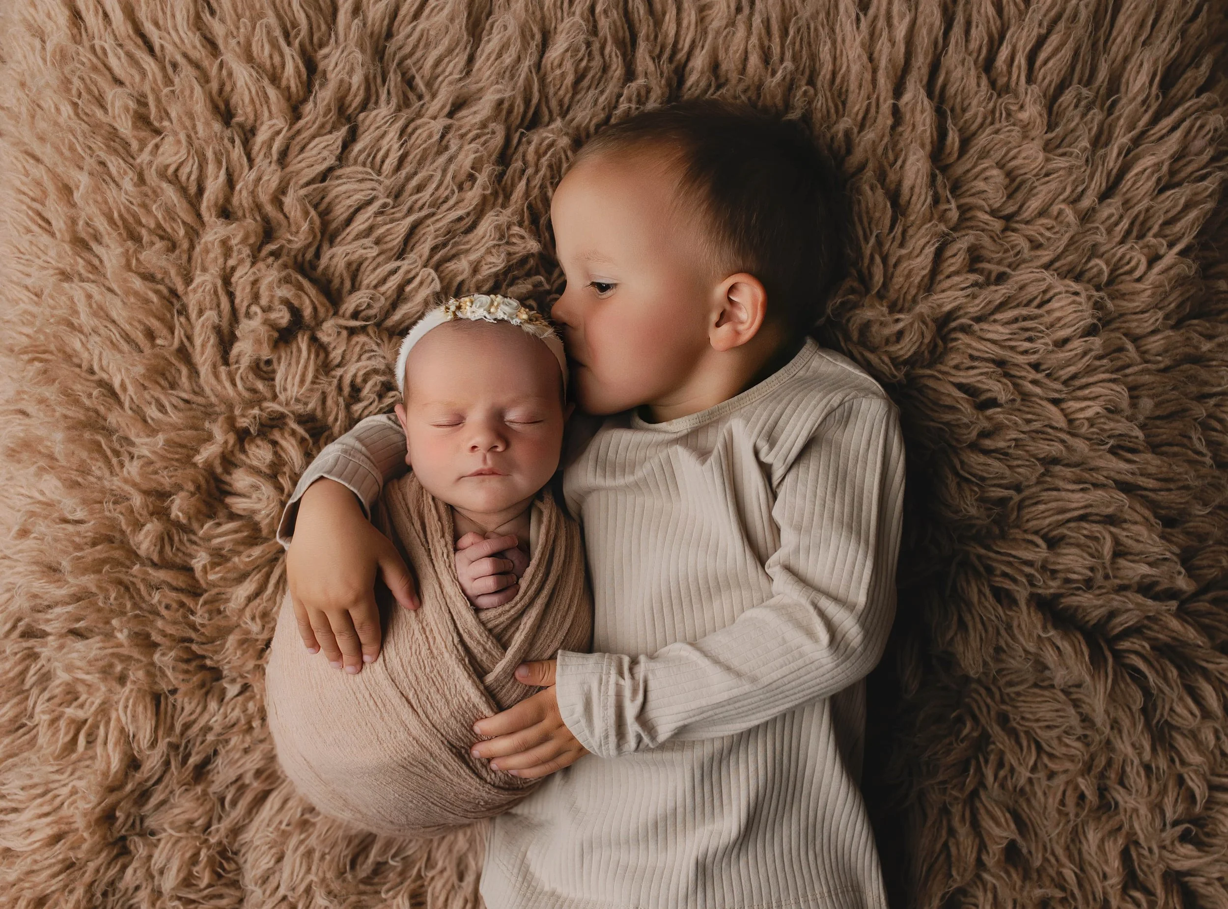 To små barn ligger sammen på en myk, lys brun pelsdekket overflate. Den eldre gutten kysser den sovende babyen på pannen.