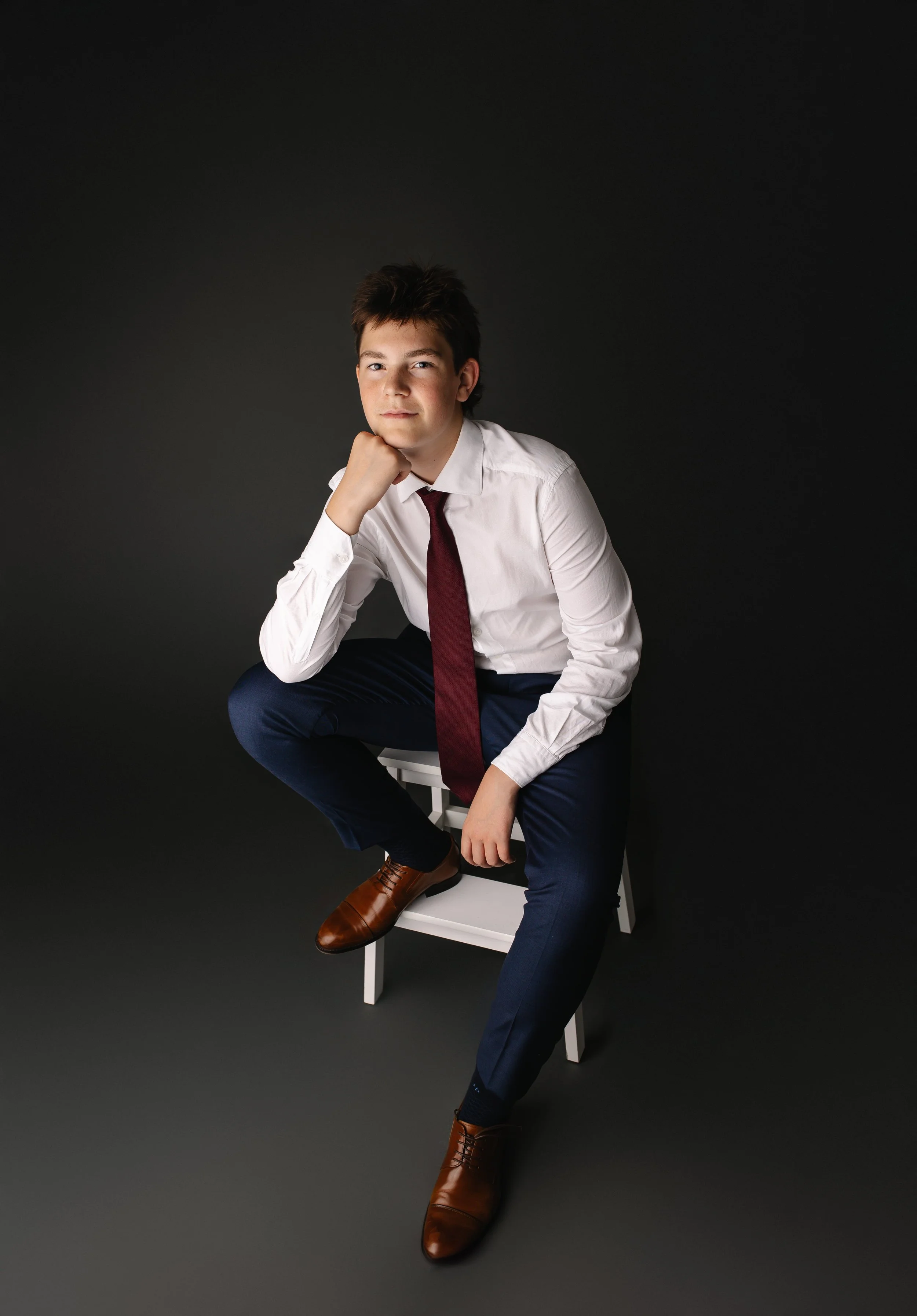 Ung mann i hvit skjorte, mørk blå bukser, og brune sko, sitter på en hvit stol mot svart bakgrunn, ser direkte i kamera.