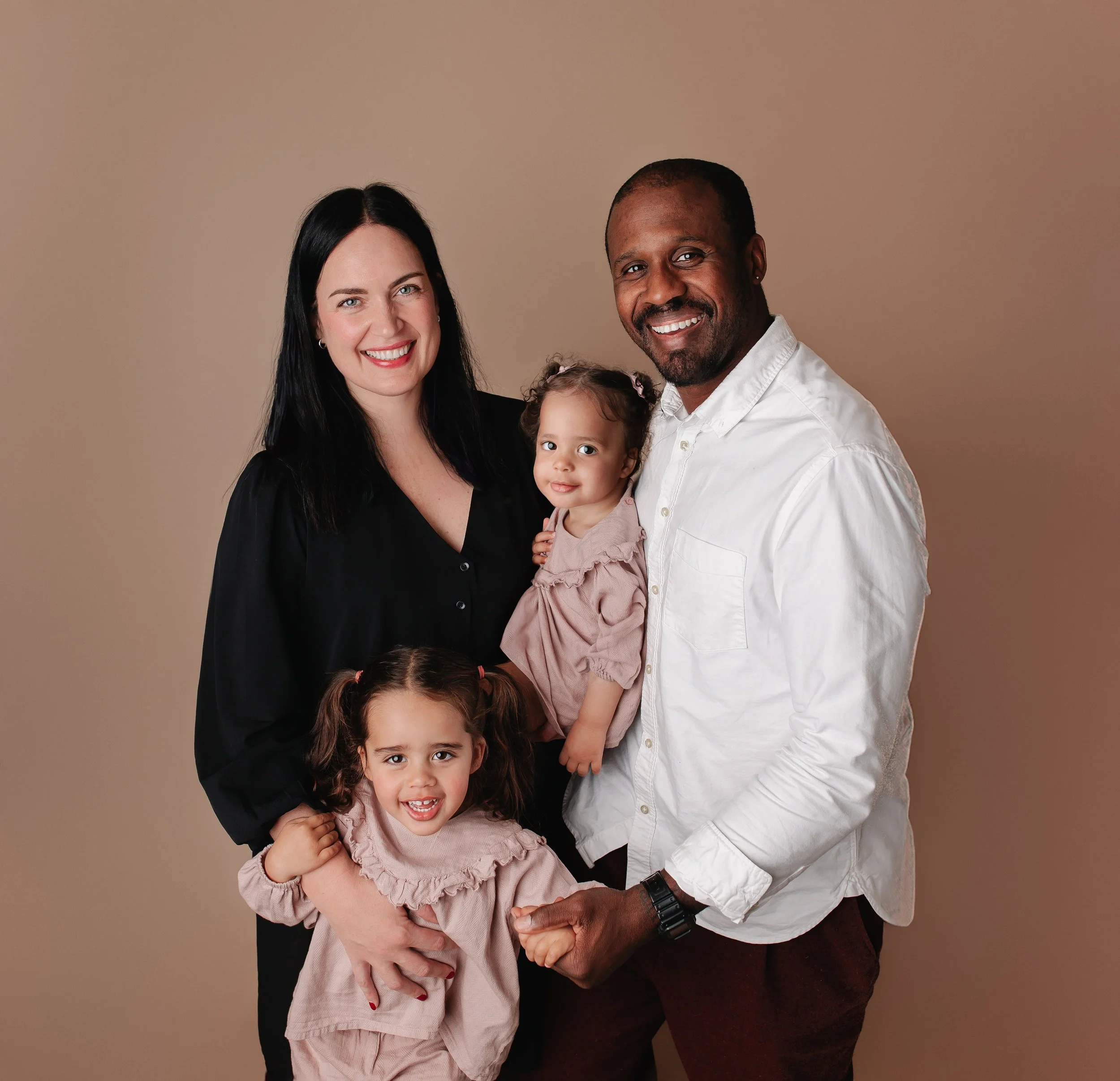 Et smilende familie med to voksne og to unge jenter står foran en beige bakgrunn.