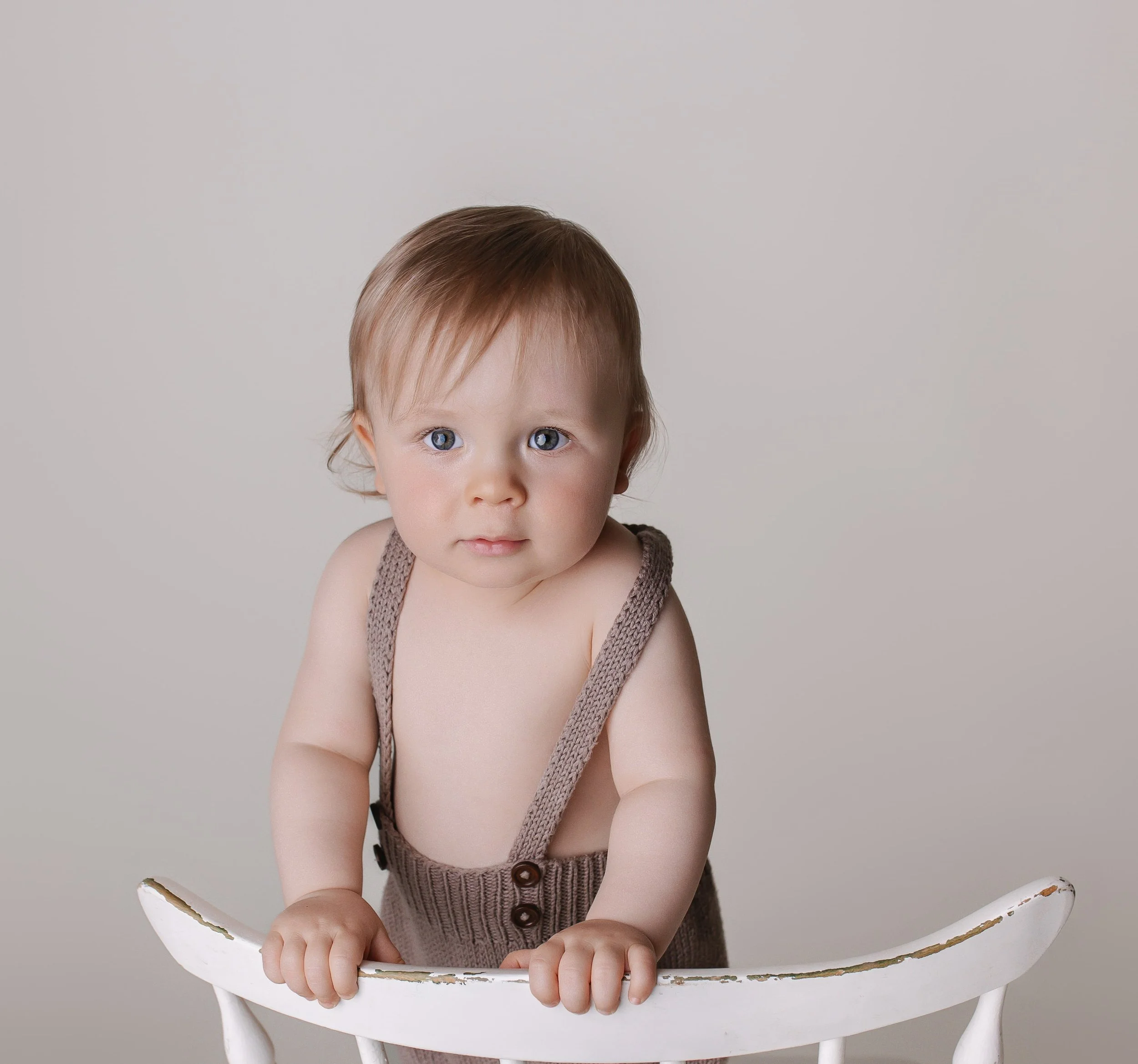 En baby med blå øyne og lyst brunt hår står oppreist i en hvit stol, iført beige rompertøy med skulderstropper.