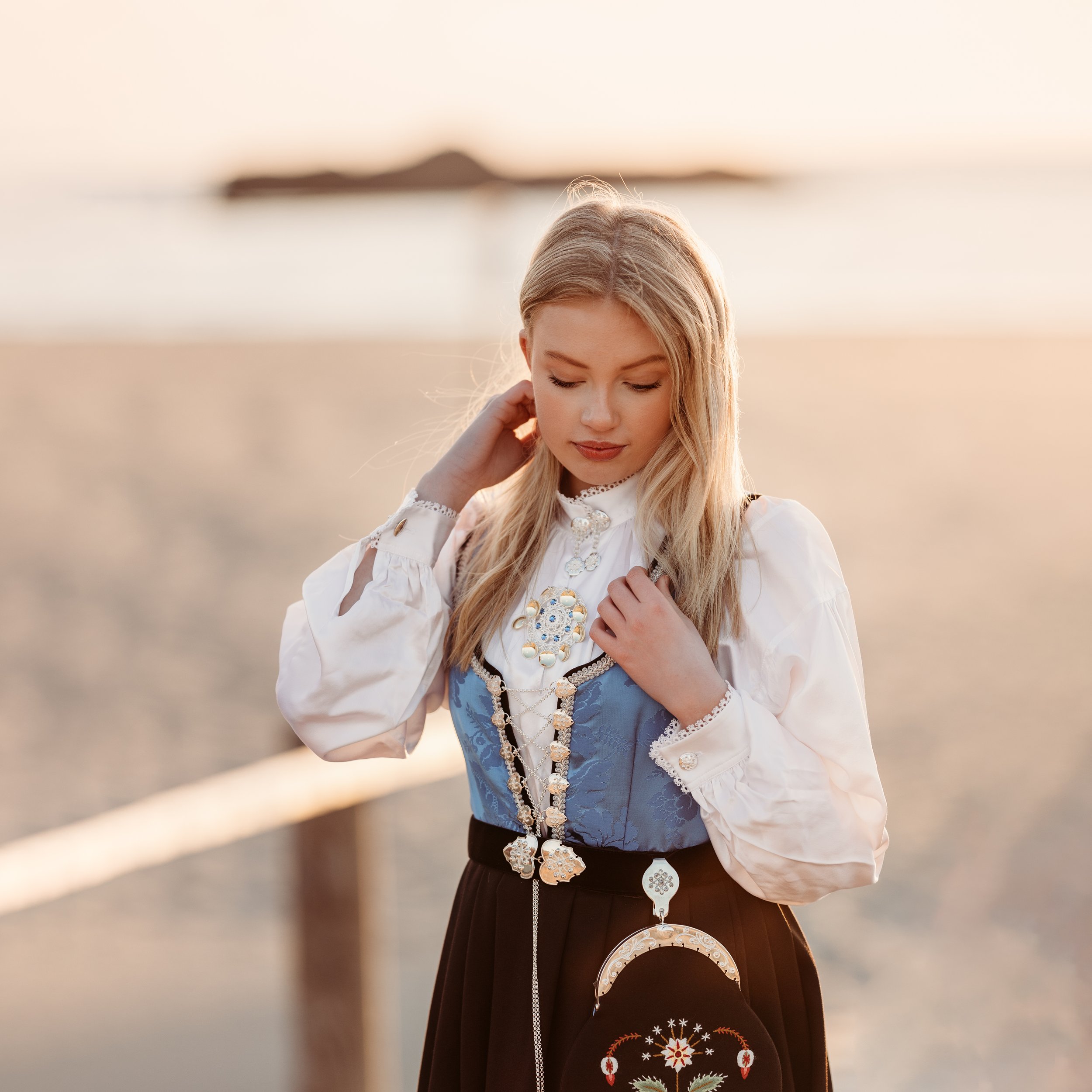 Ung kvinne i tradisjonell norsk bunad med fjær i håret, står på en strand under solnedgang.