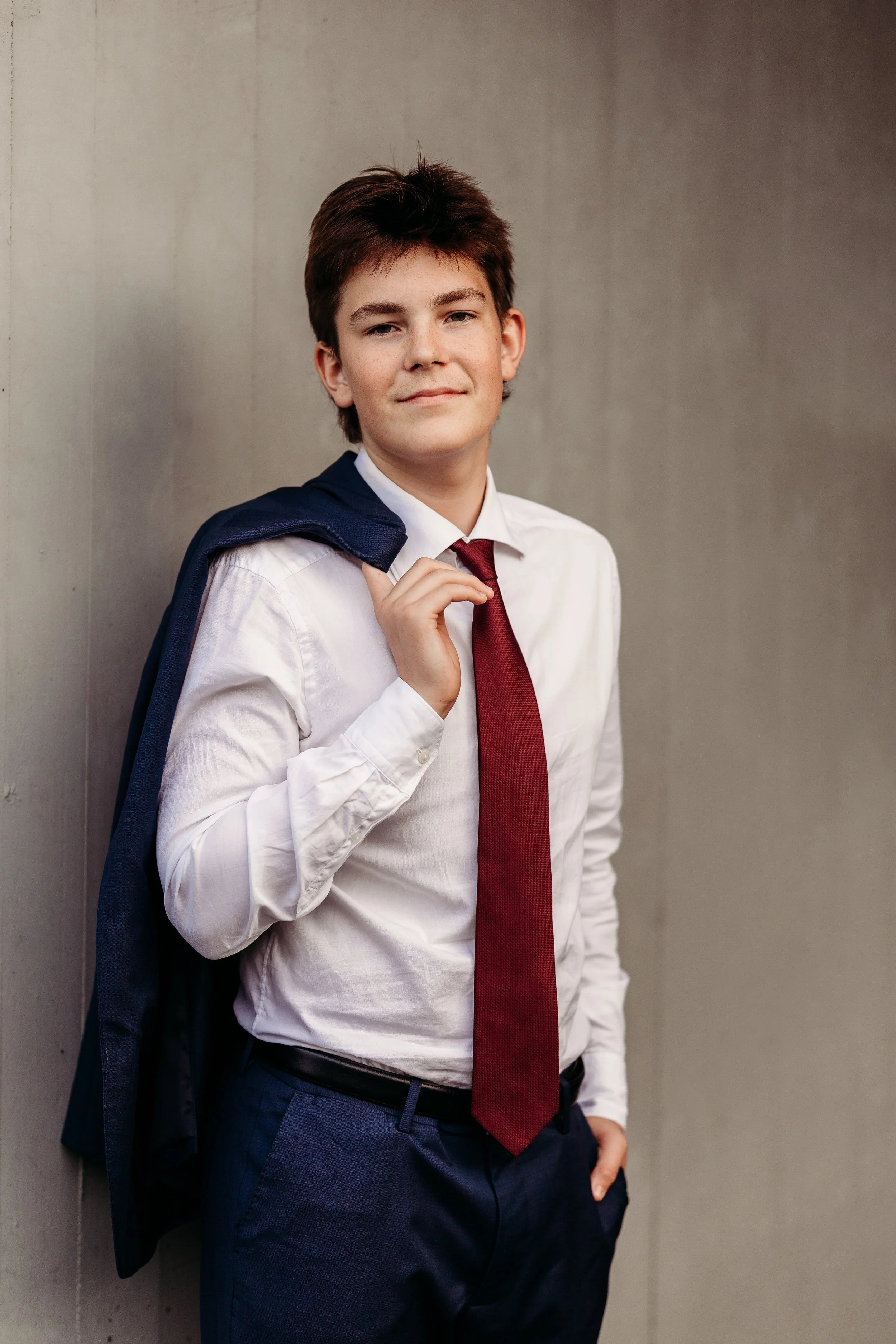 Ung mann i dress, hvit skjorte og rød slips, som bærer en mørk dressjakke over skulderen, står mot en grå vegg.