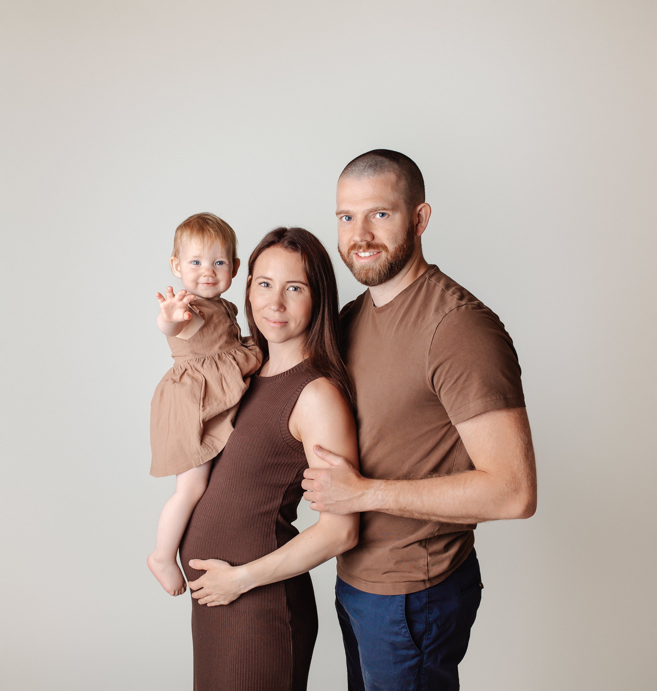 Et gravidt par står sammen med deres lille datter mot en lys, enkel bakgrunn.