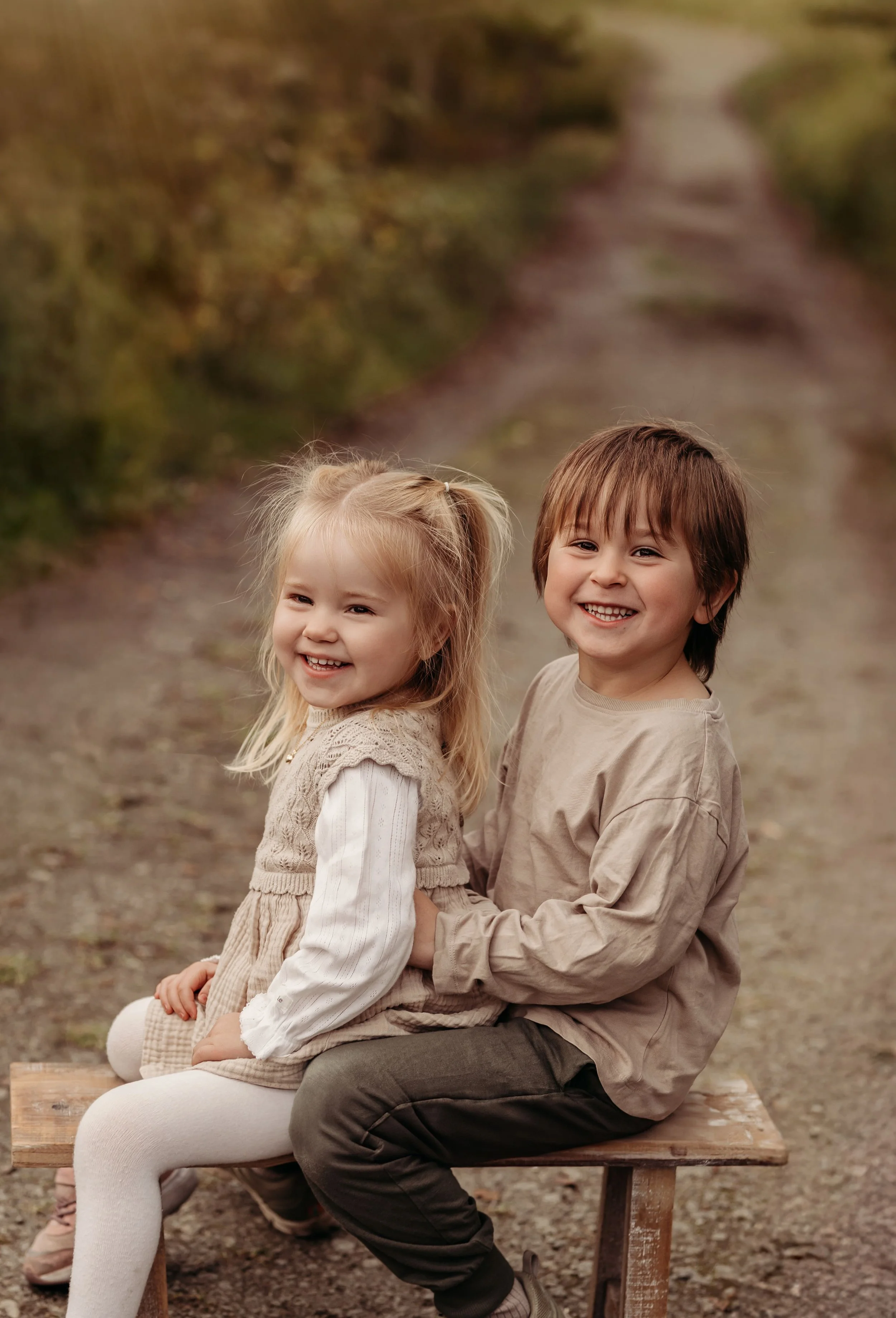 To barn sitter på en trebord utendørs, smiler og ler, omgitt av natur med en rolig sti i bakgrunnen.