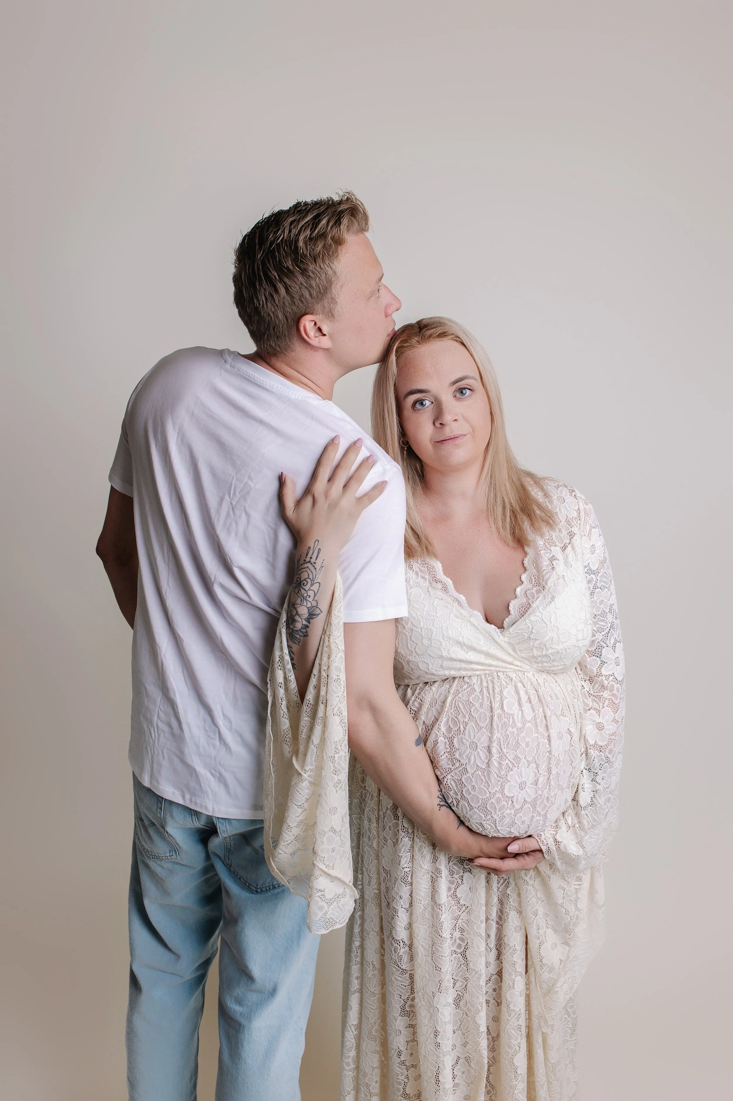 En gravid kvinne i en lys blondekjole holder magen og står foran en mann som kysser hennes hode, mot en enkel lys bakgrunn.