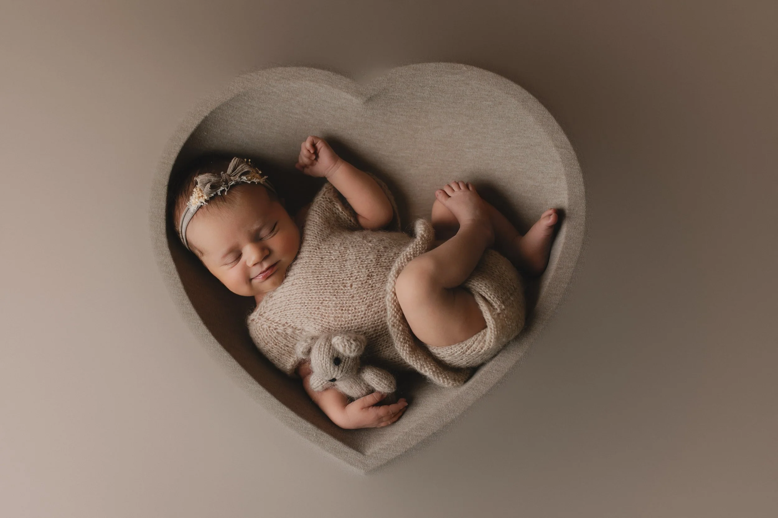 Søvnende baby i en hjerteformet seng, med en liten bamse i hånden, iført en strikket drakt og en hodebånd.