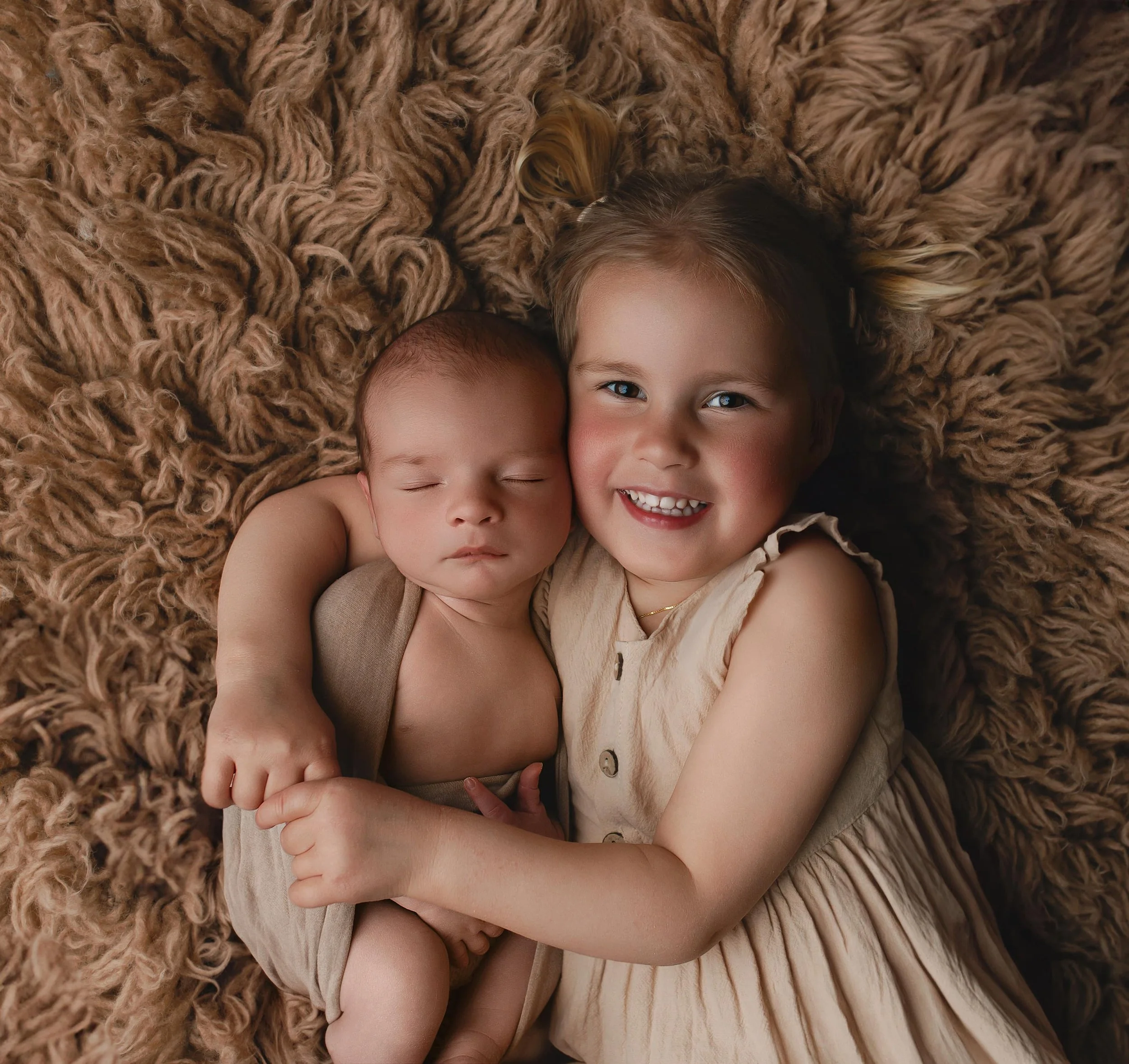 En liten jente holder en sovende baby, begge ligger på en myk, brun pelsaktig teppe. Jenta smiler bredt og har krøllete hår.