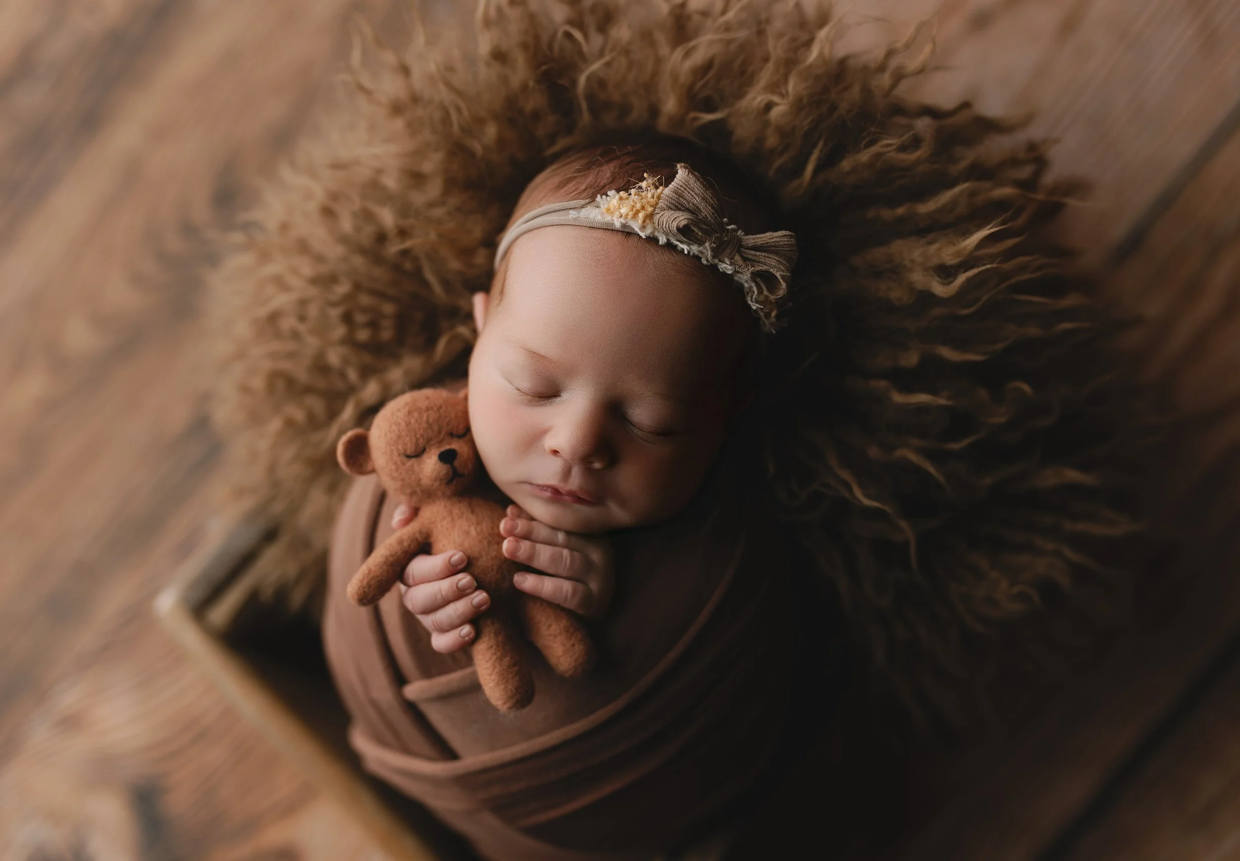 Et sovende spedbarn med et lyserosa plagg og en elastisk hodebånd med blomst, som holder en liten brunt plysteddy, liggende på en rund, brun pels og en tregulvflate.