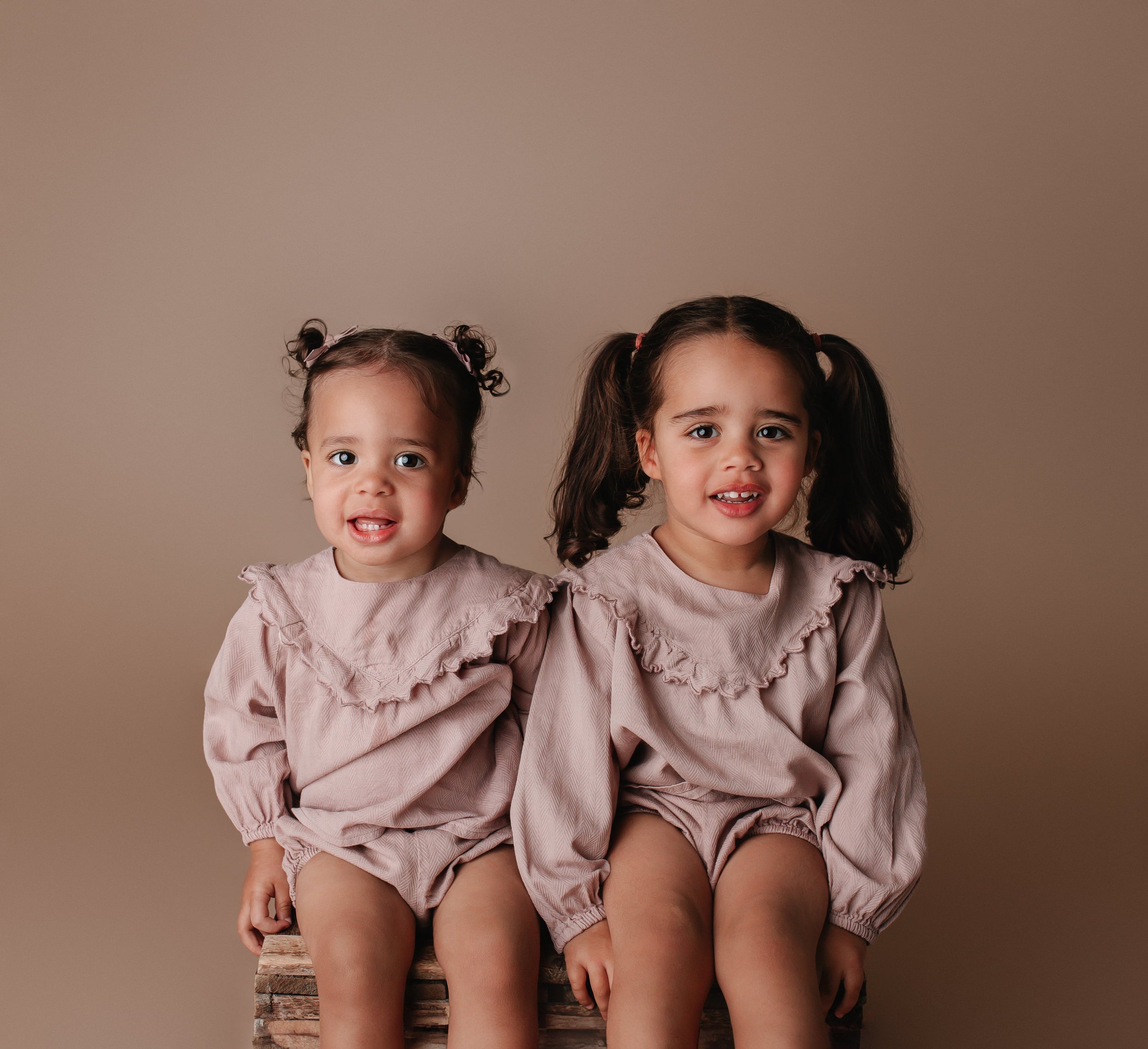 To unge jenter med mørkt hår og løse klær sitter på en trestokk mot en beige bakgrunn.