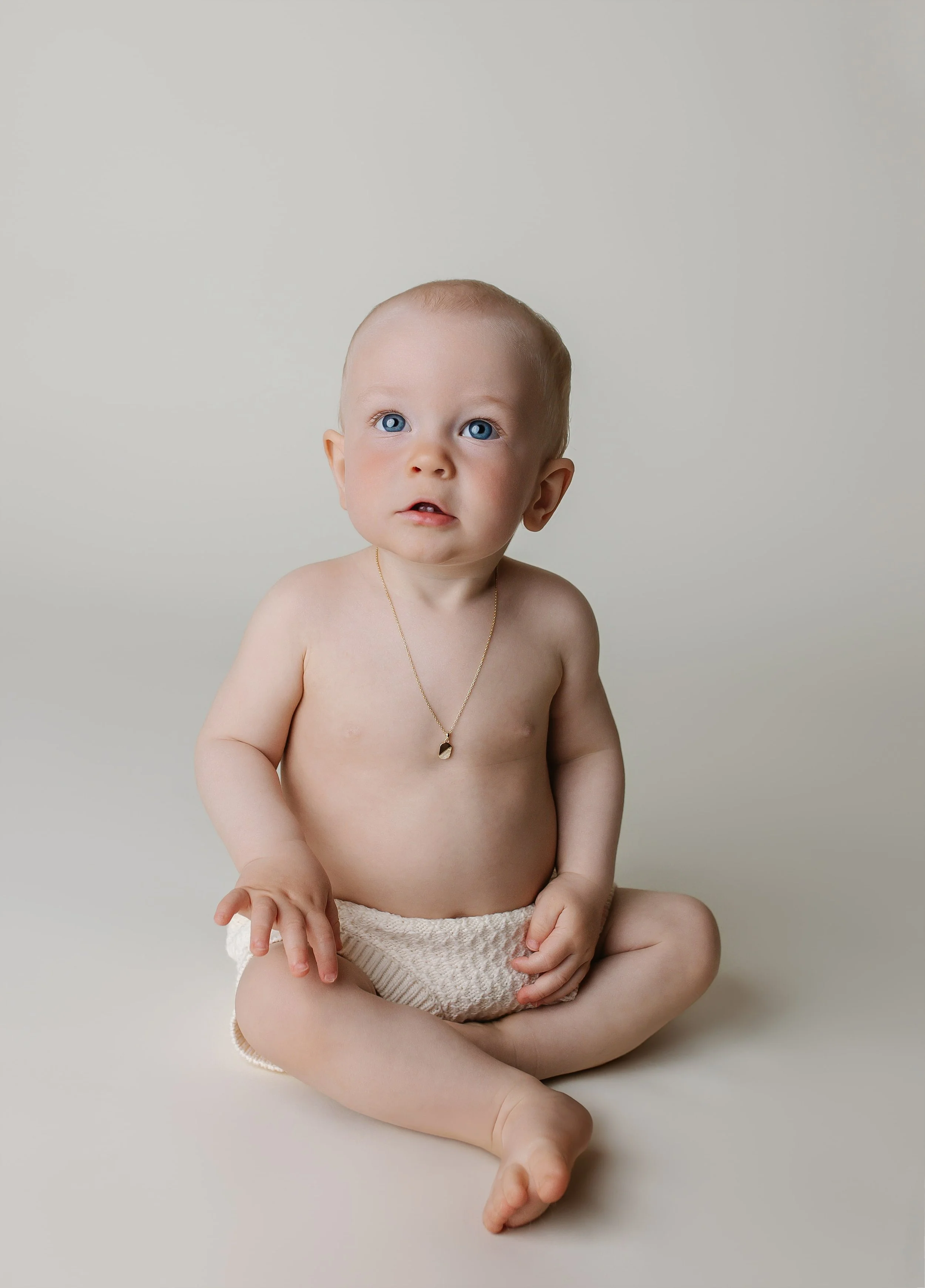 En guttunge sitter på gulvet uten klær, iført en armknapp og en halskjede, med blå øyne og lys hud, mot en lys bakgrunn.
