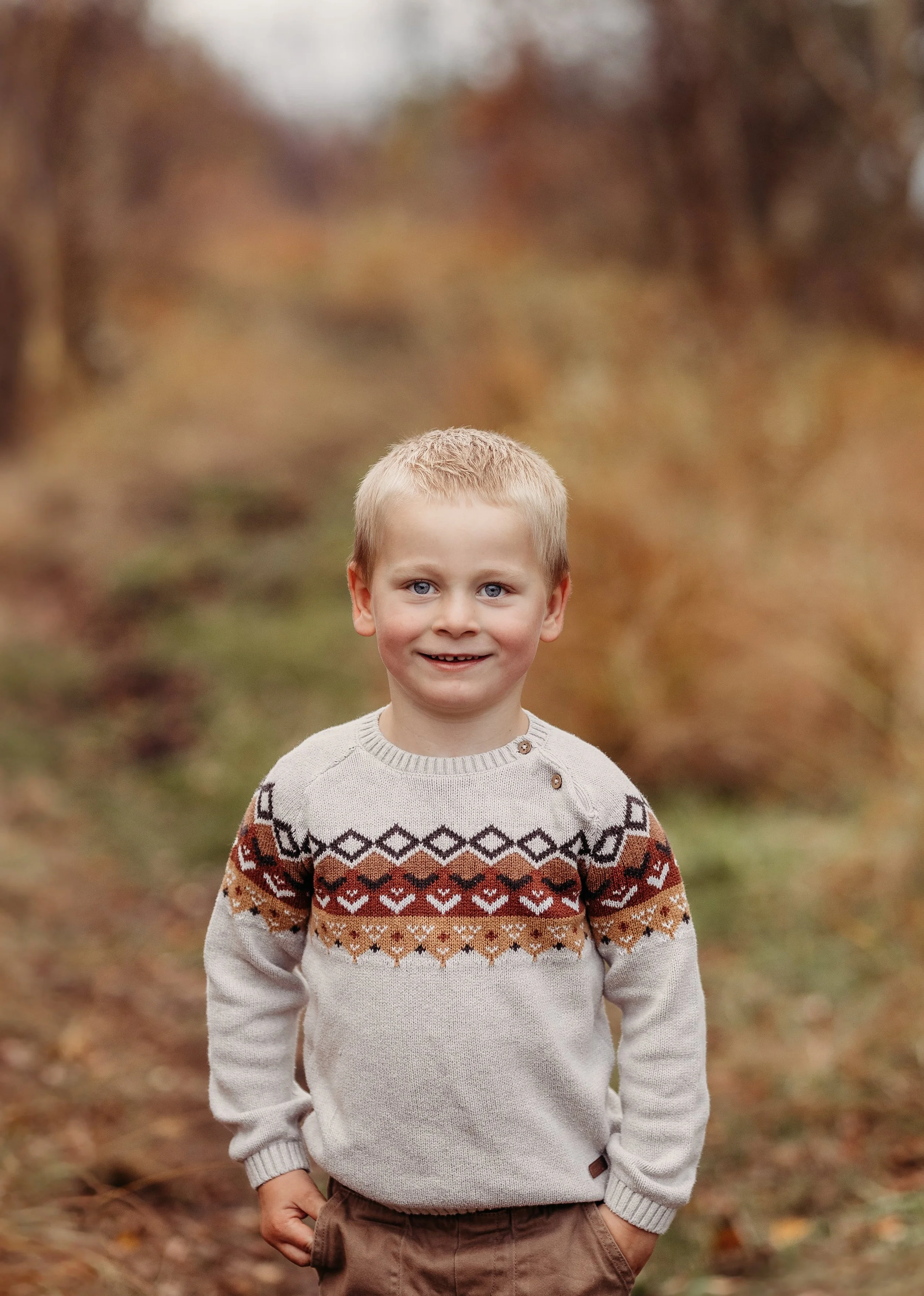 Ung gutt med blond hår og blå øyne, smilende i en park med høstfarger på trærne, iført en lys genser med mønster og brune bukser.