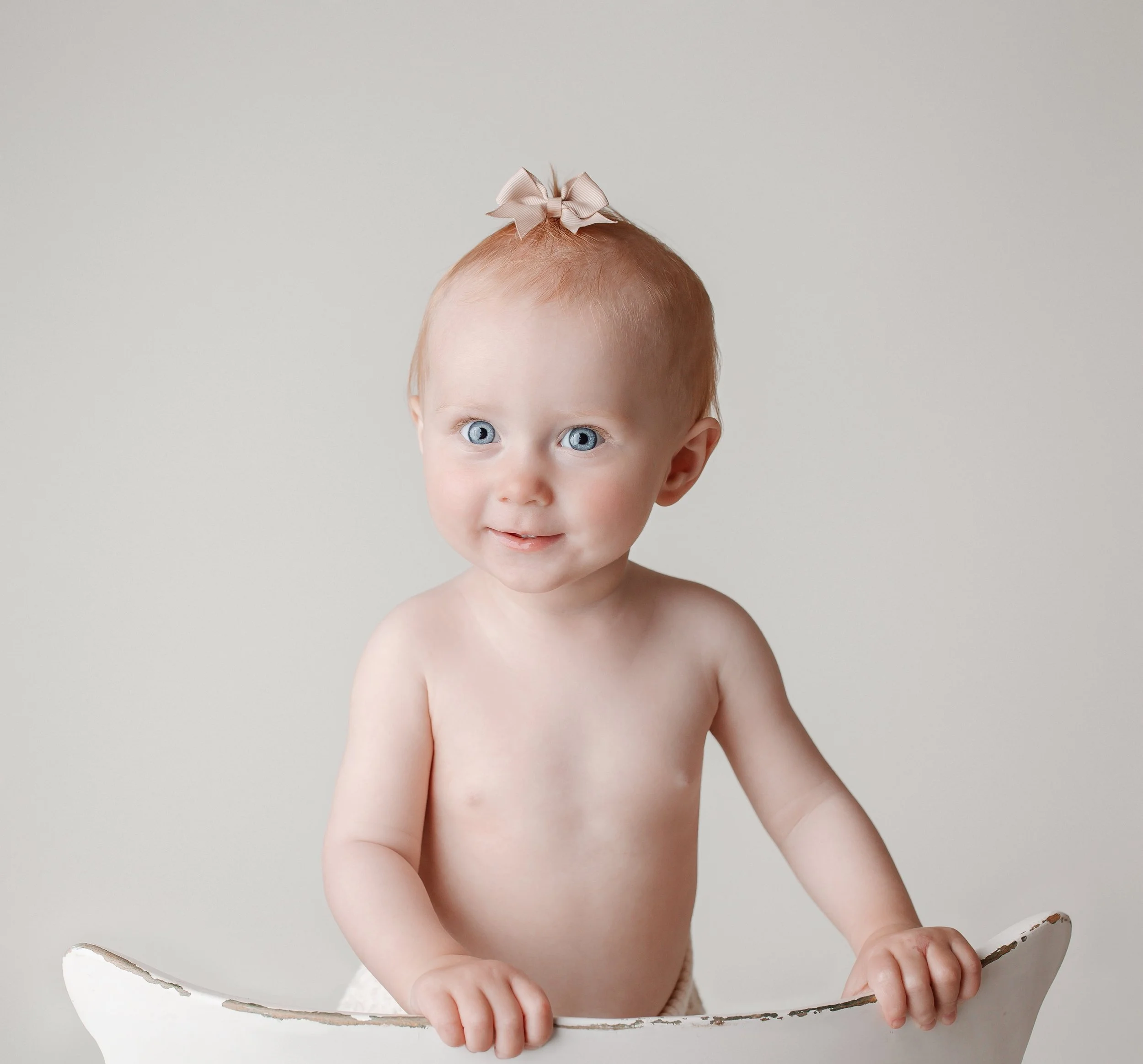 En blid babyjente med blå øyne, uten klær, med en beige sløyfe i håret, holder i en hvit gjenstand, mot en enkel lysgrå bakgrunn.