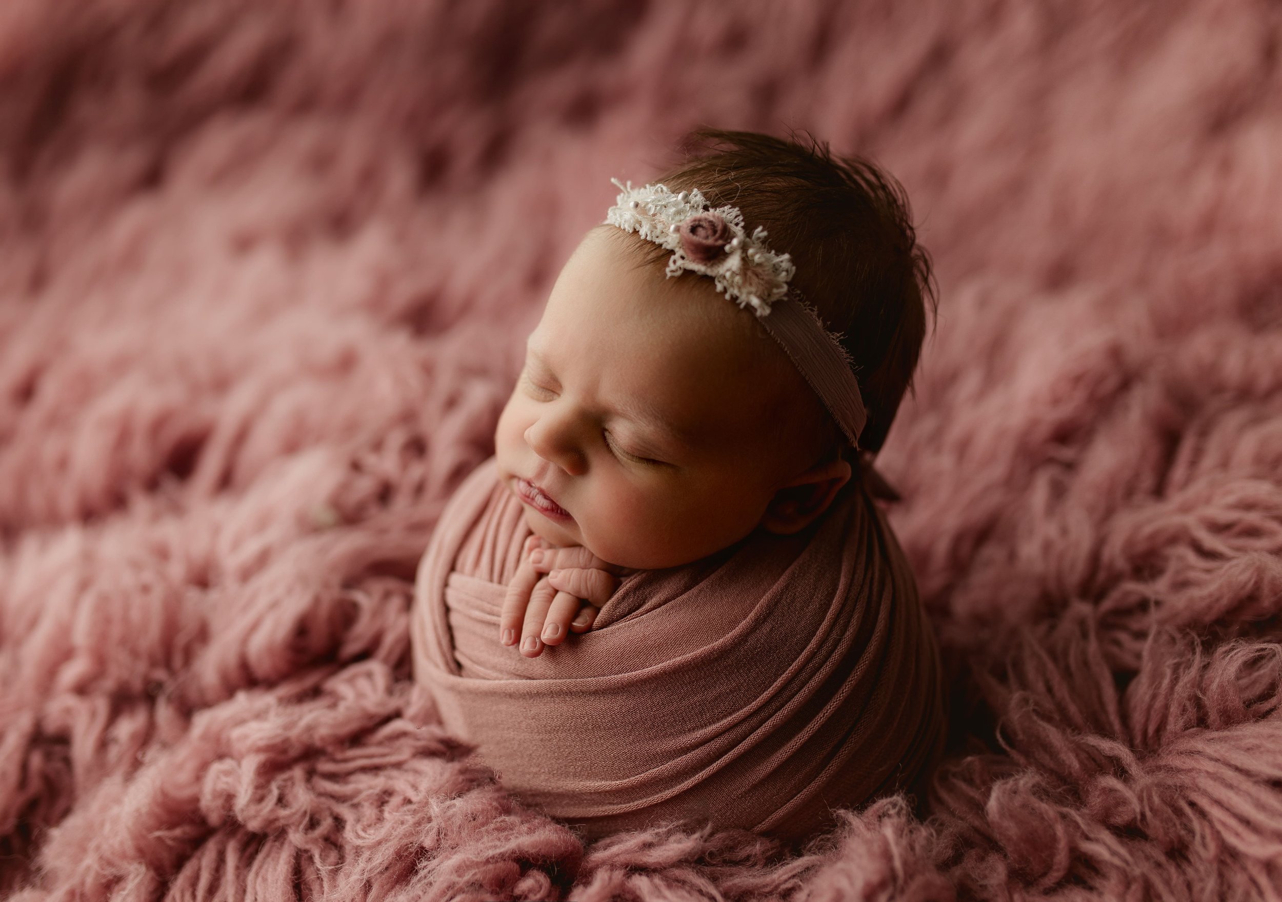 Søt nyfødt baby med blomstertopp, liggende på rosa fluffy teppe, med lukkede øyne og et smil.