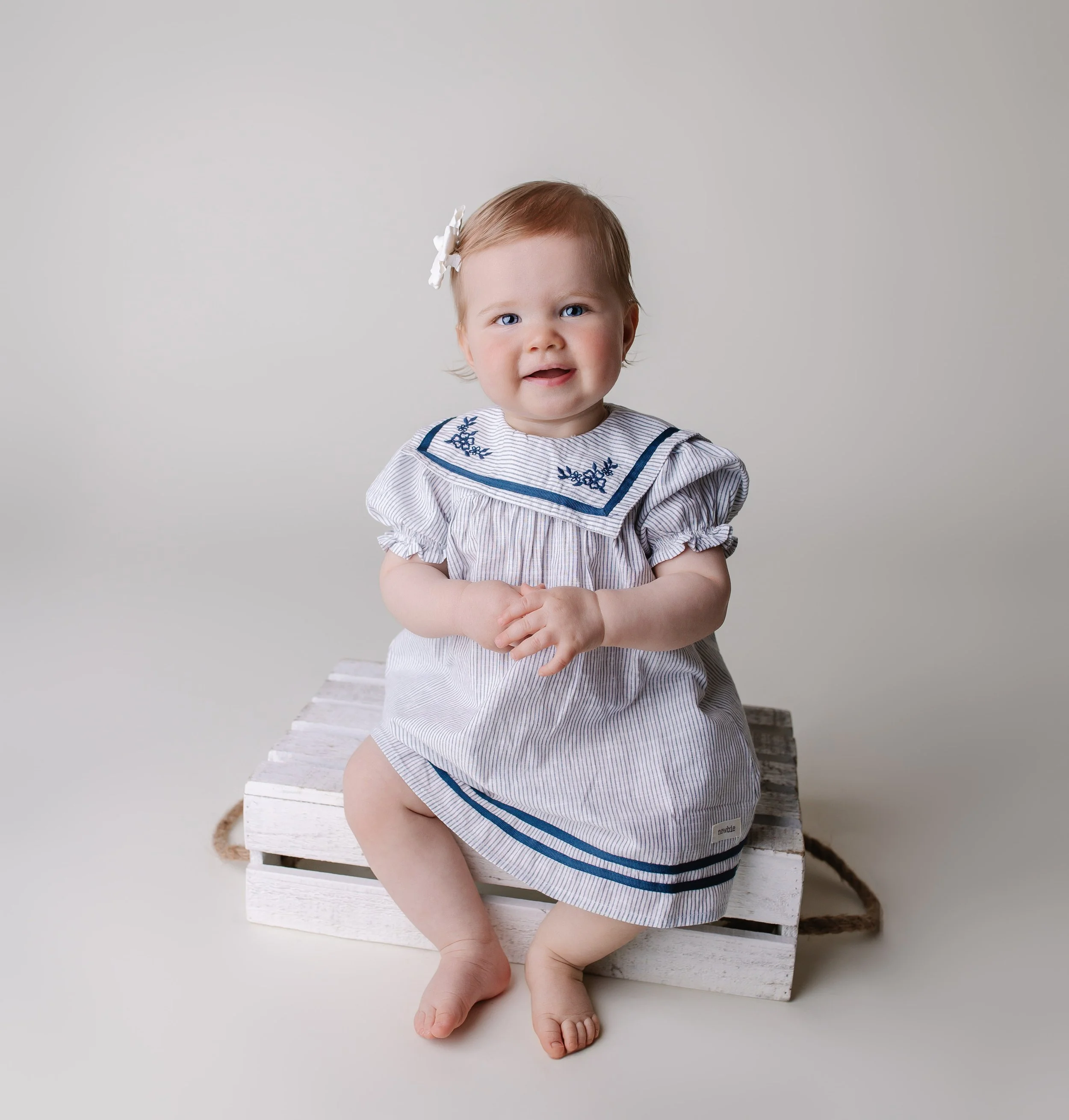 En liten jente med blond hår, kledd i en striper kjole med blå detaljer, sitter på en hvit tre kasse mot en lys grå bakgrunn, smiler og holder hendene sammen.