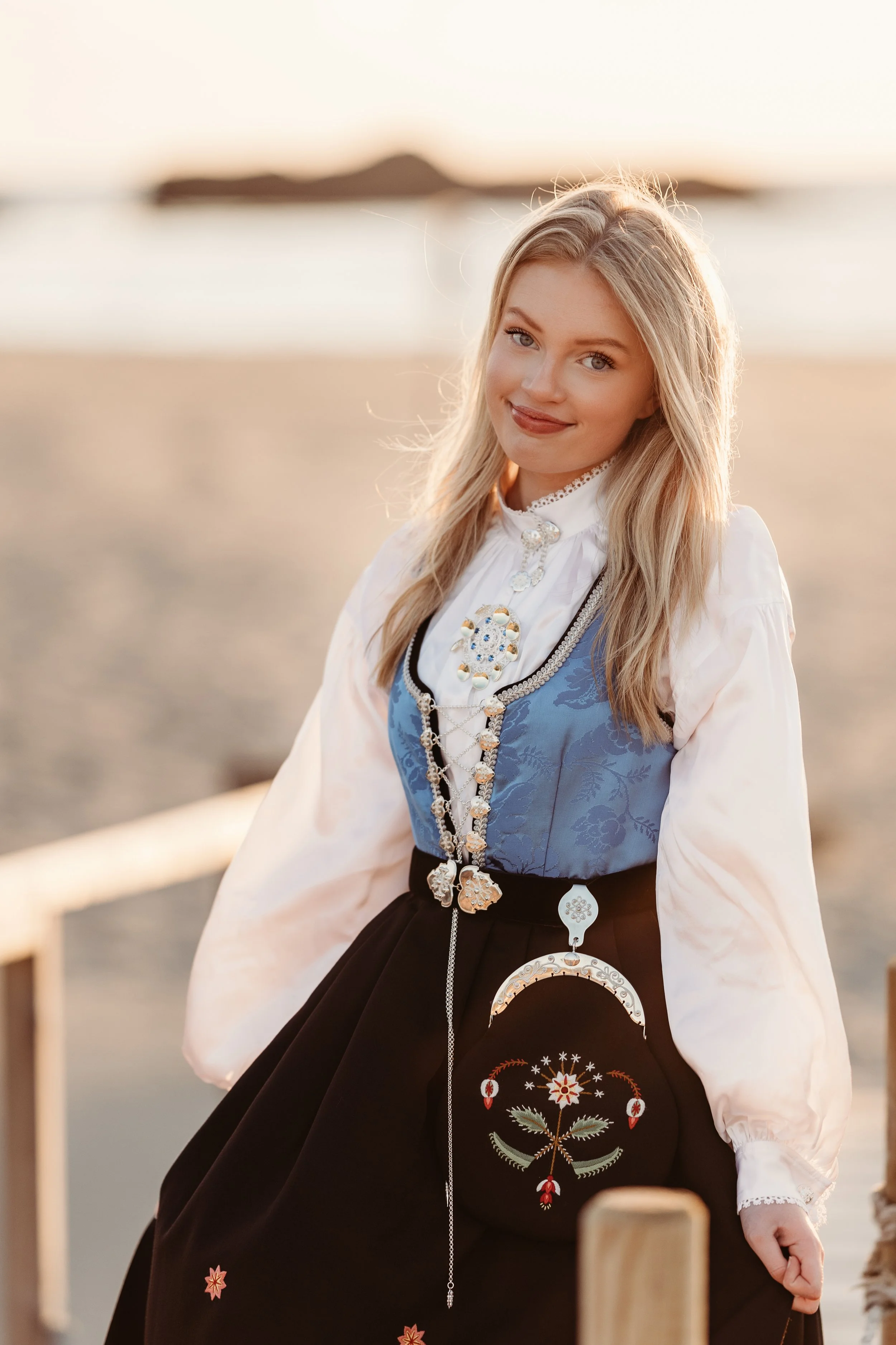 En ung kvinne i tradisjonell norsk bunad står ved sjøen under solnedgangen.