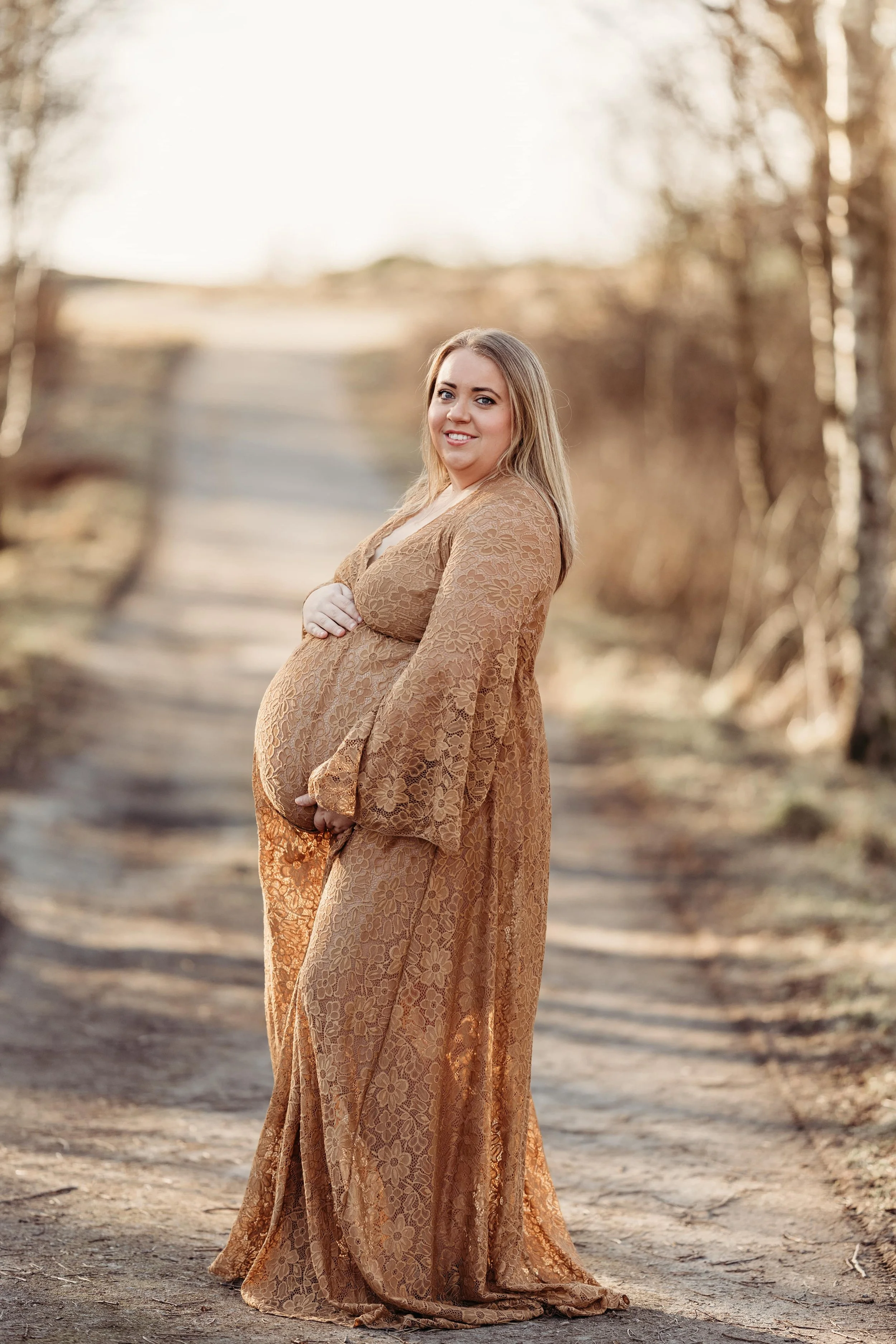 En mengder gravid kvinne i en brun, blomstrete kjole, som står på en skogssti med trær i bakgrunnen. Hun smiler og holder hånden på magen.