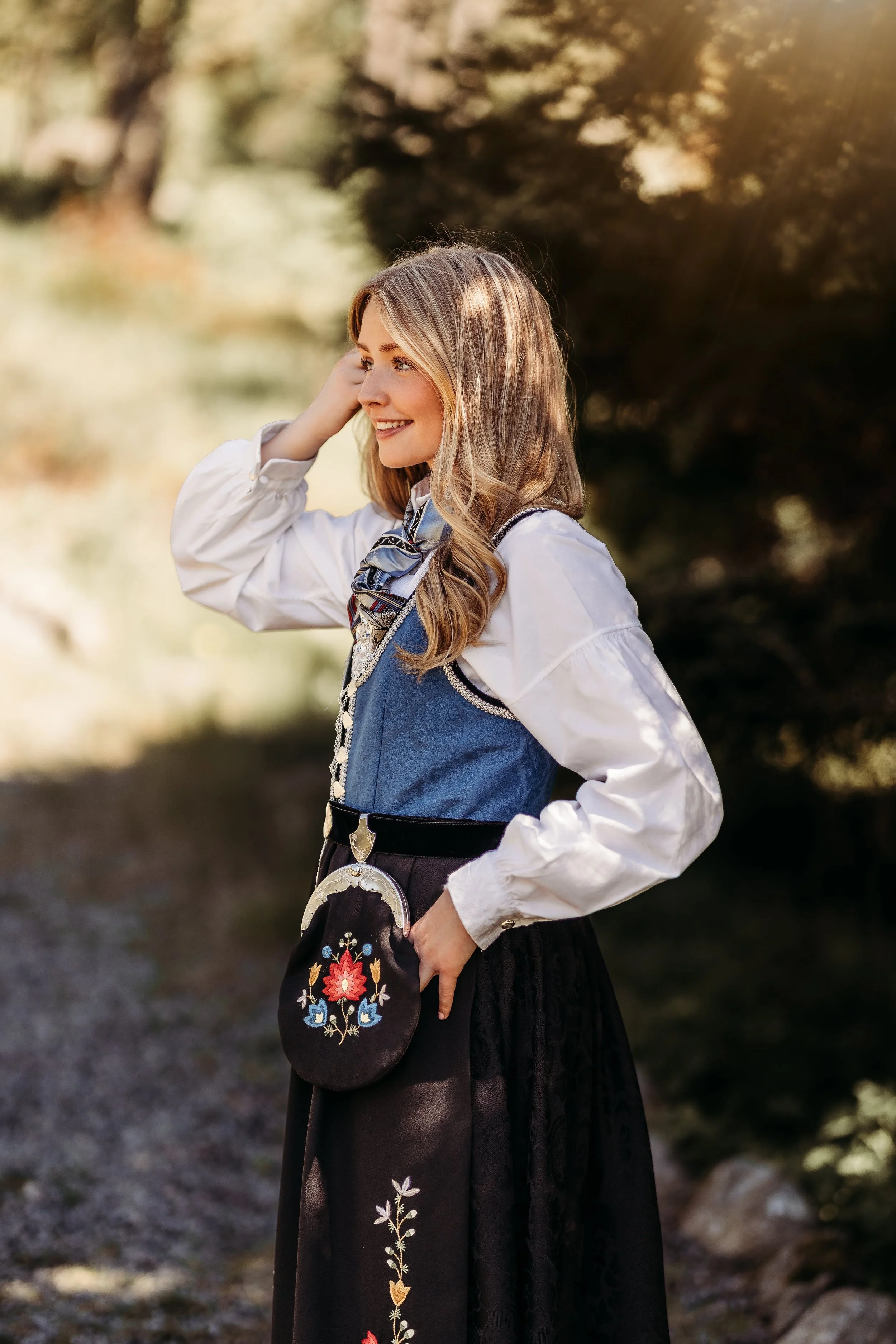 Kvinne i tradisjonell norsk bunad smilende, utendørs naturbakgrunn.