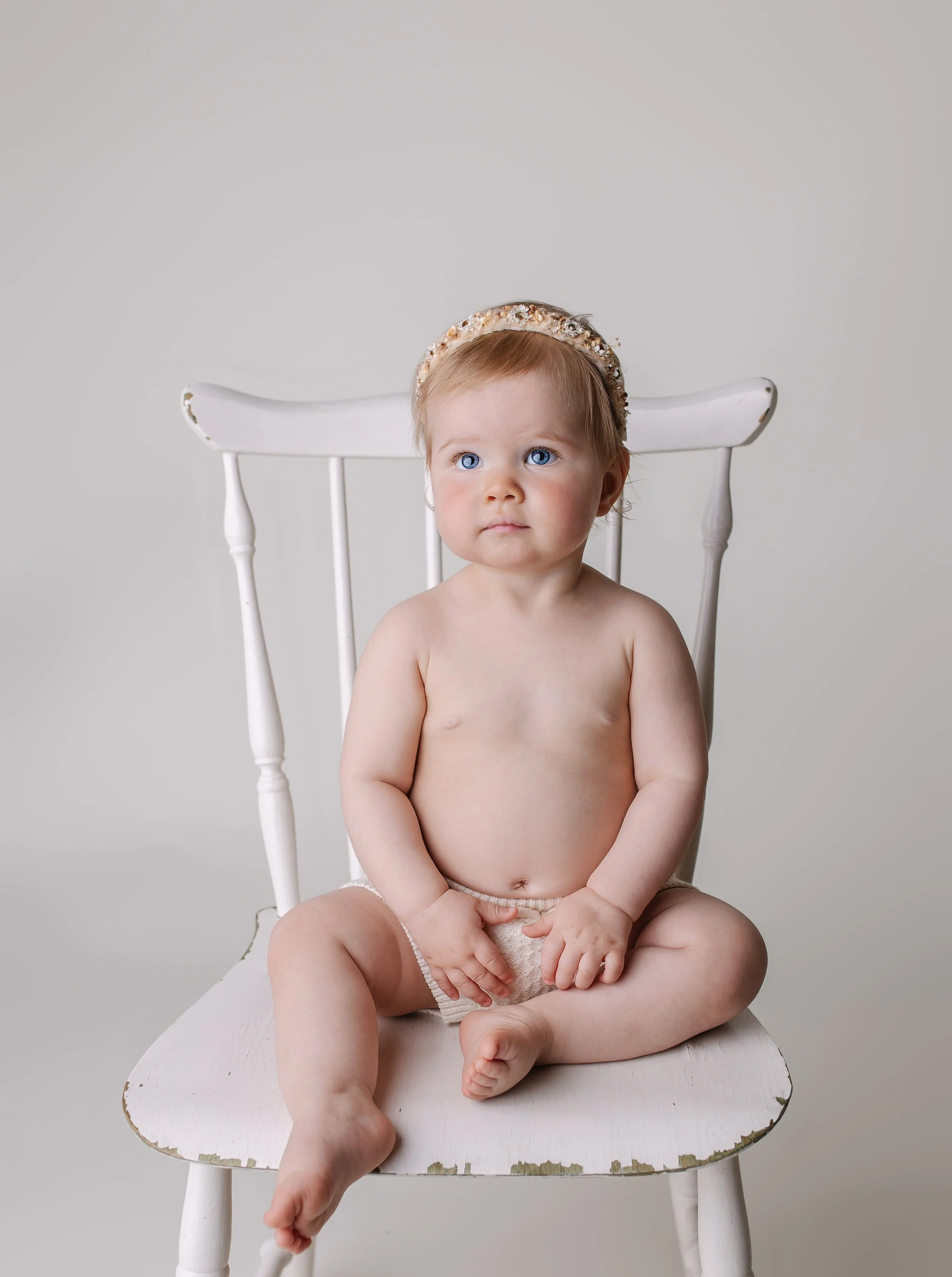 En liten barndr og uten klær som sitter på en hvit trone med en blomsterhodebånd.