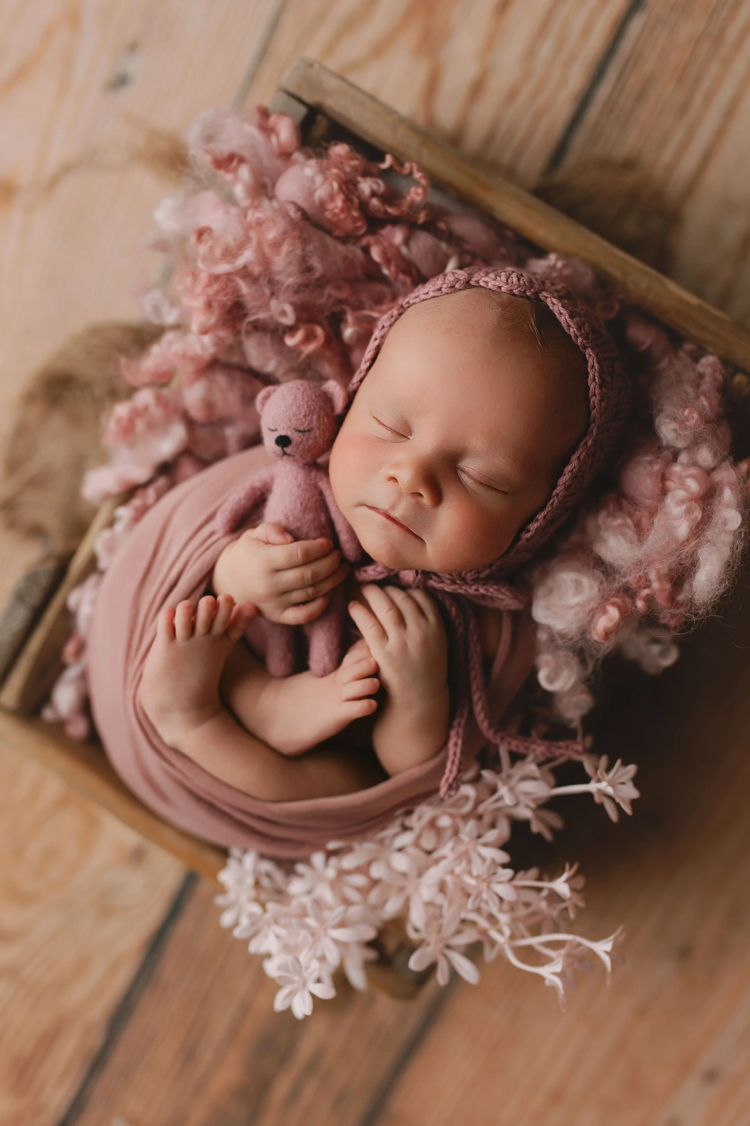 En sovende baby kledd i rosa, holdende en liten rosa bamse, omgitt av rosa og hvite ullfibre, lagt i en trekasse på et treverk.
