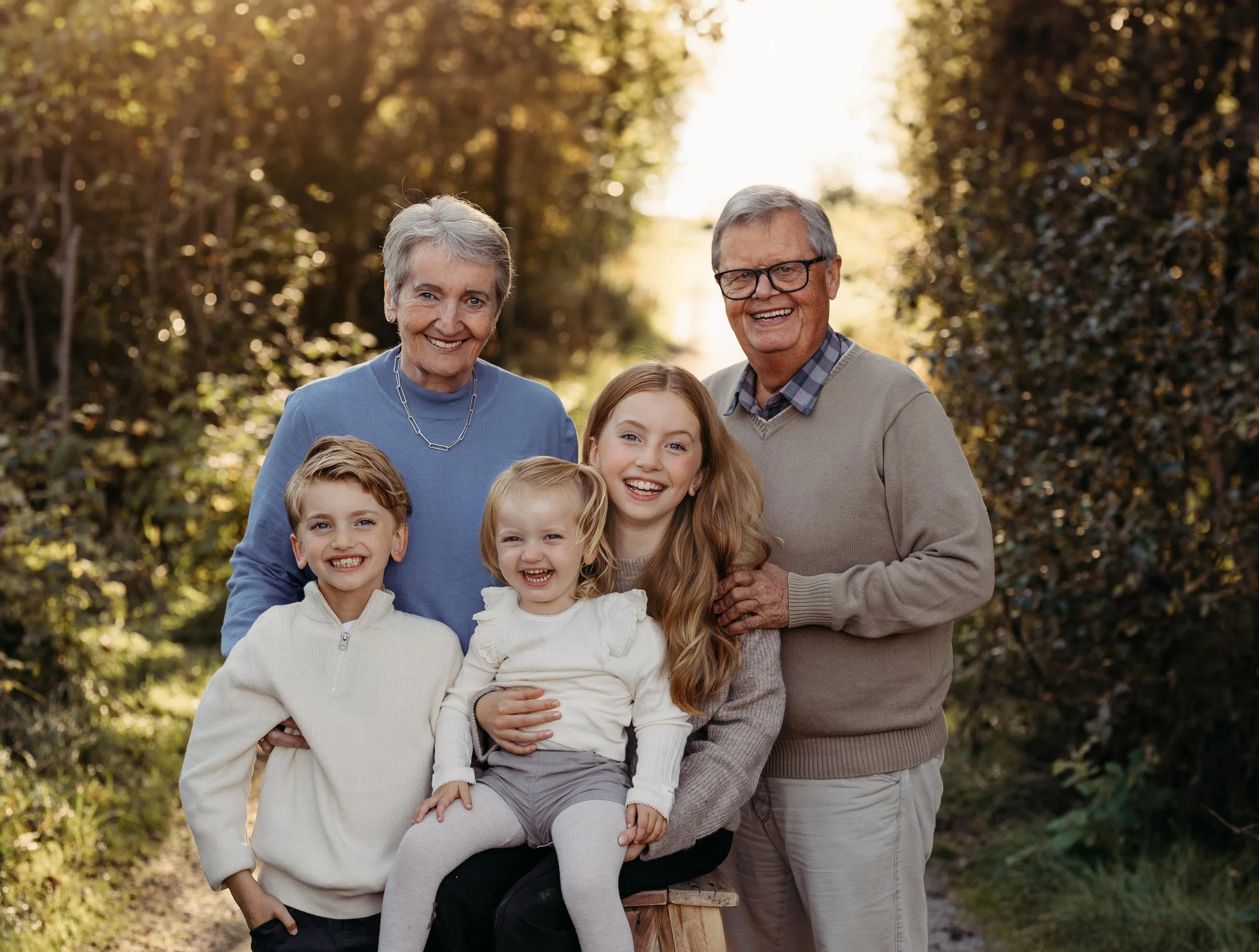 Bilde av en gammel familie samlet utendørs, smilende, under trær med høstfarger.