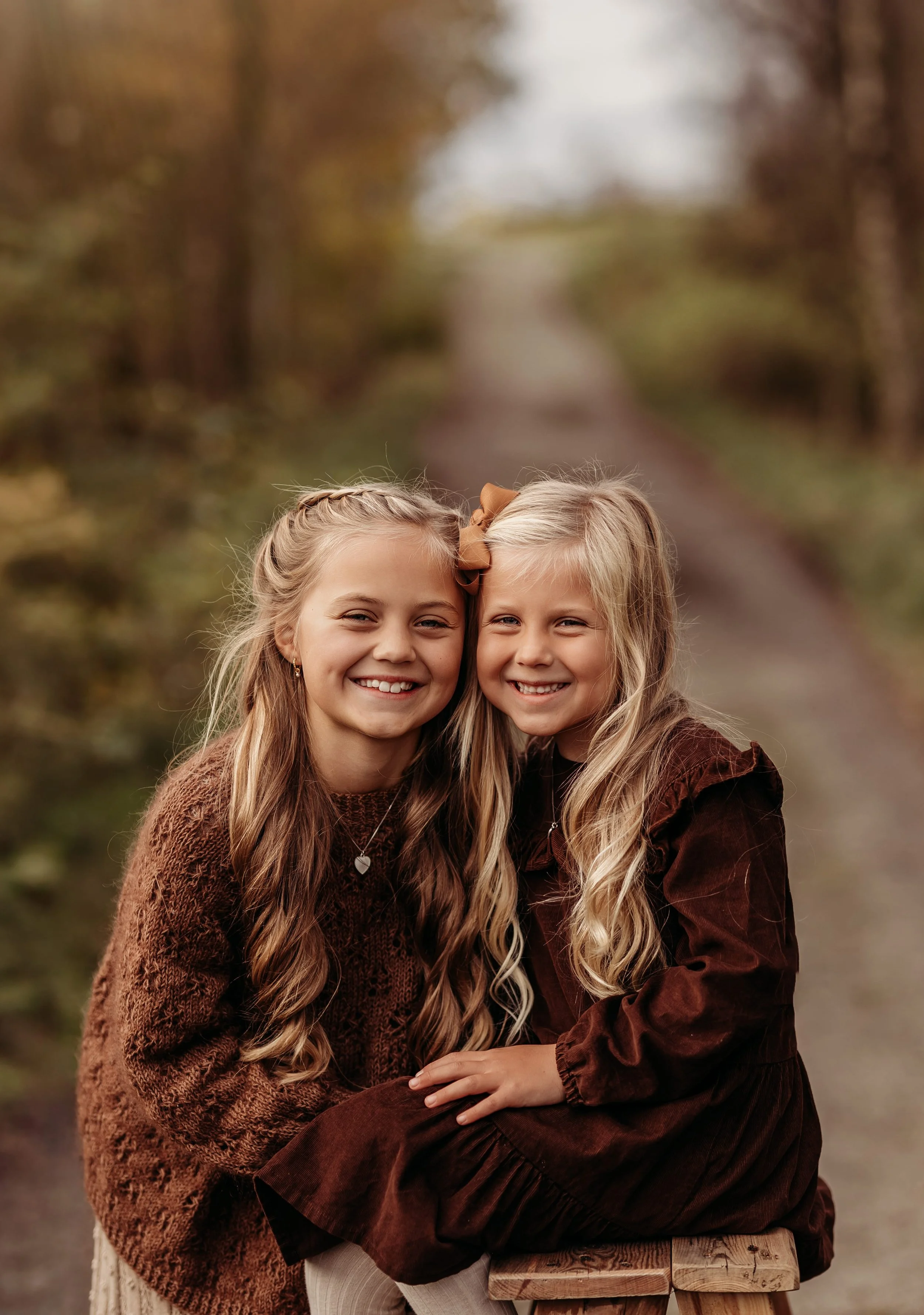 To unge jenter som smiler og sitter sammen utendørs i en skog stien, høstfarger i bakgrunnen.