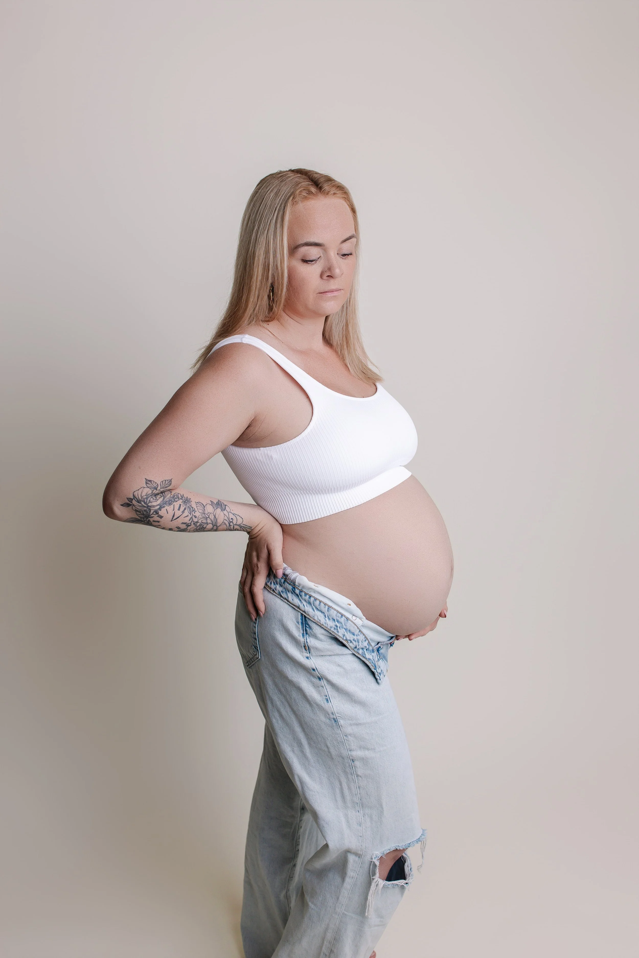 En gravid kvinne med blondt hår i hvit trenings- eller tank-topp, løstsittende jeans med hull, står mot en enkel lys bakgrunn.