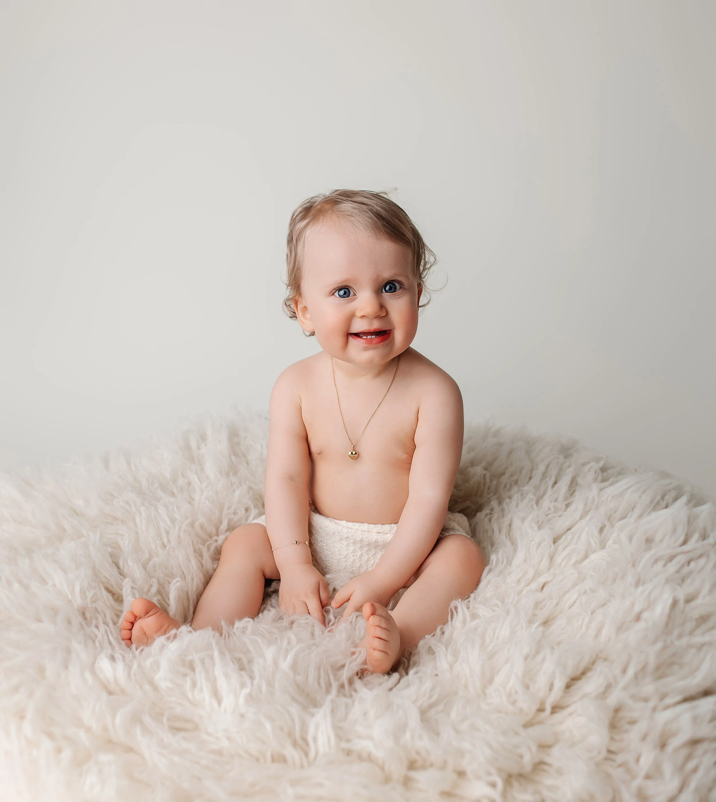 En baby med blå øyne som sitter på en myk, kremfarget pels, uten klær, med smil og halskjede, mot en lys bakgrunn.