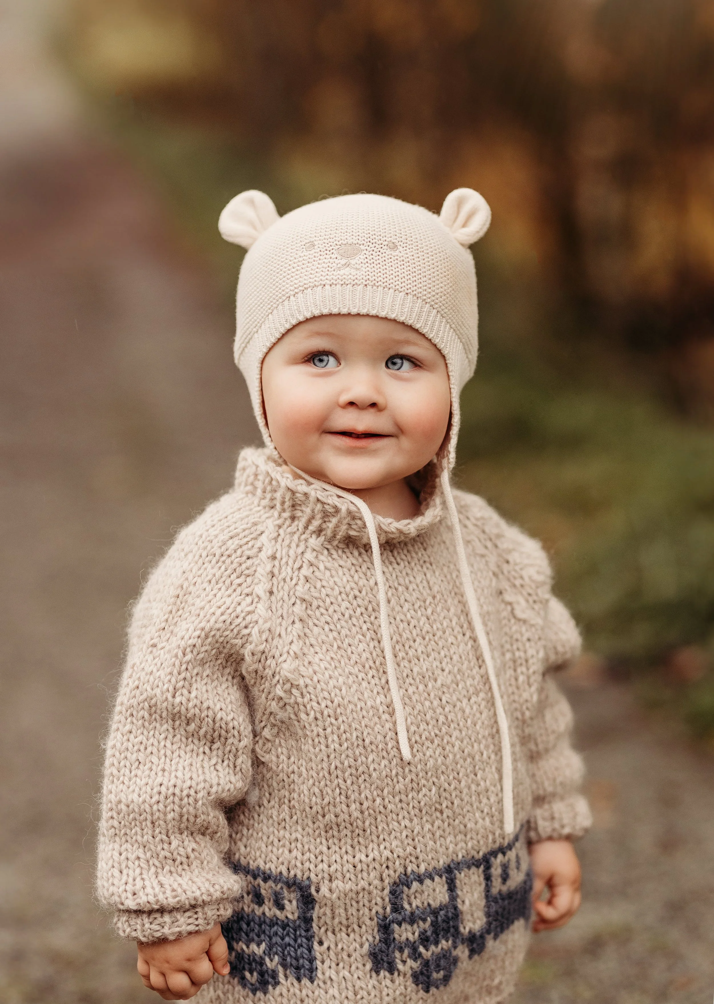 Et barnekull som går utendørs i høstlig vær, iført en beige strikkegenser og en bamse-lue. Han har blå øyne og et smil, med en bakgrunn av fargede blader.
