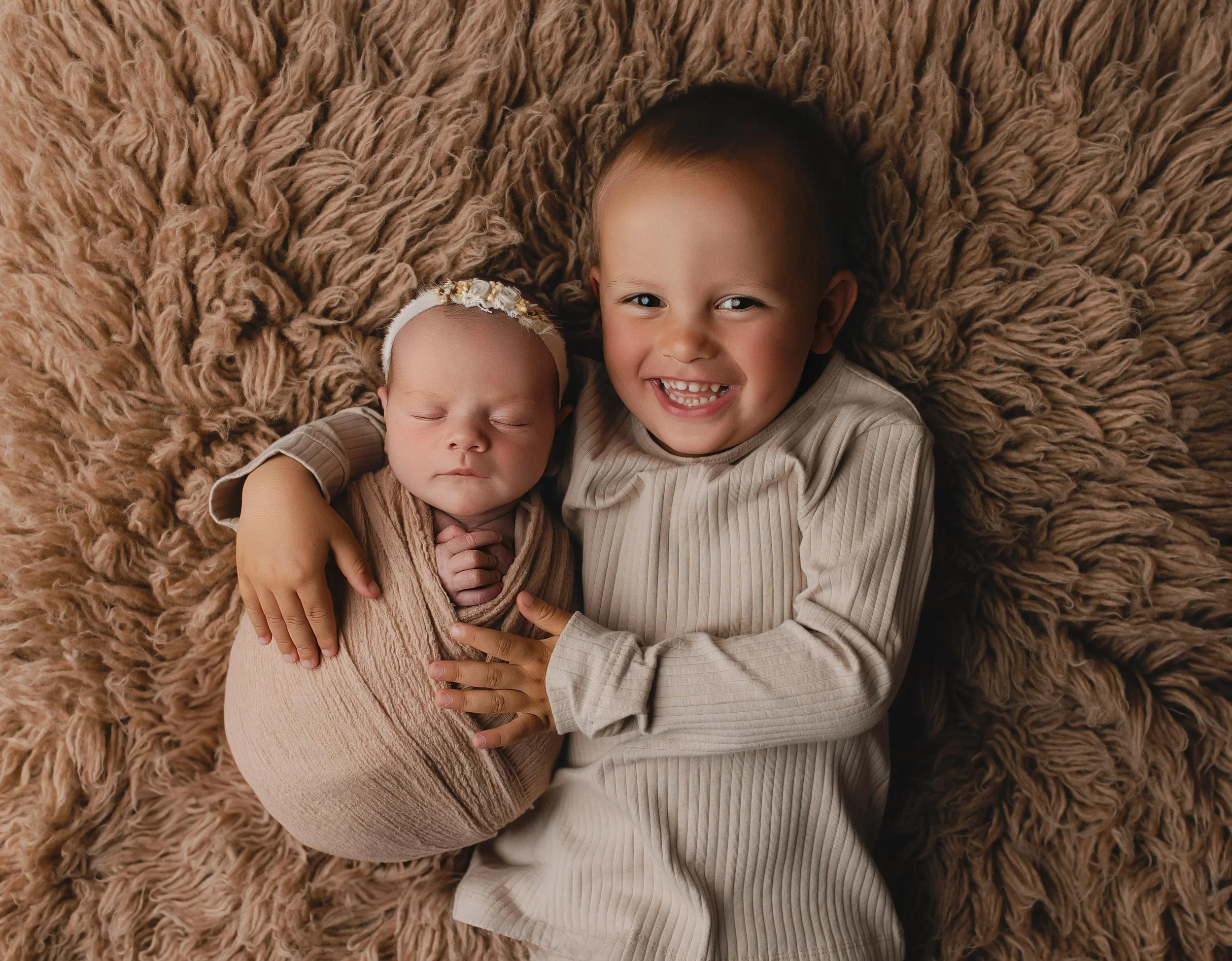 To barn ligger på en mykt, bustete, beige teppe. Den eldre gutten smiler bredt og har armen rundt den yngre babyen, som sovner; den yngre babyen har på seg en beige wrap og en blomsterkrans.