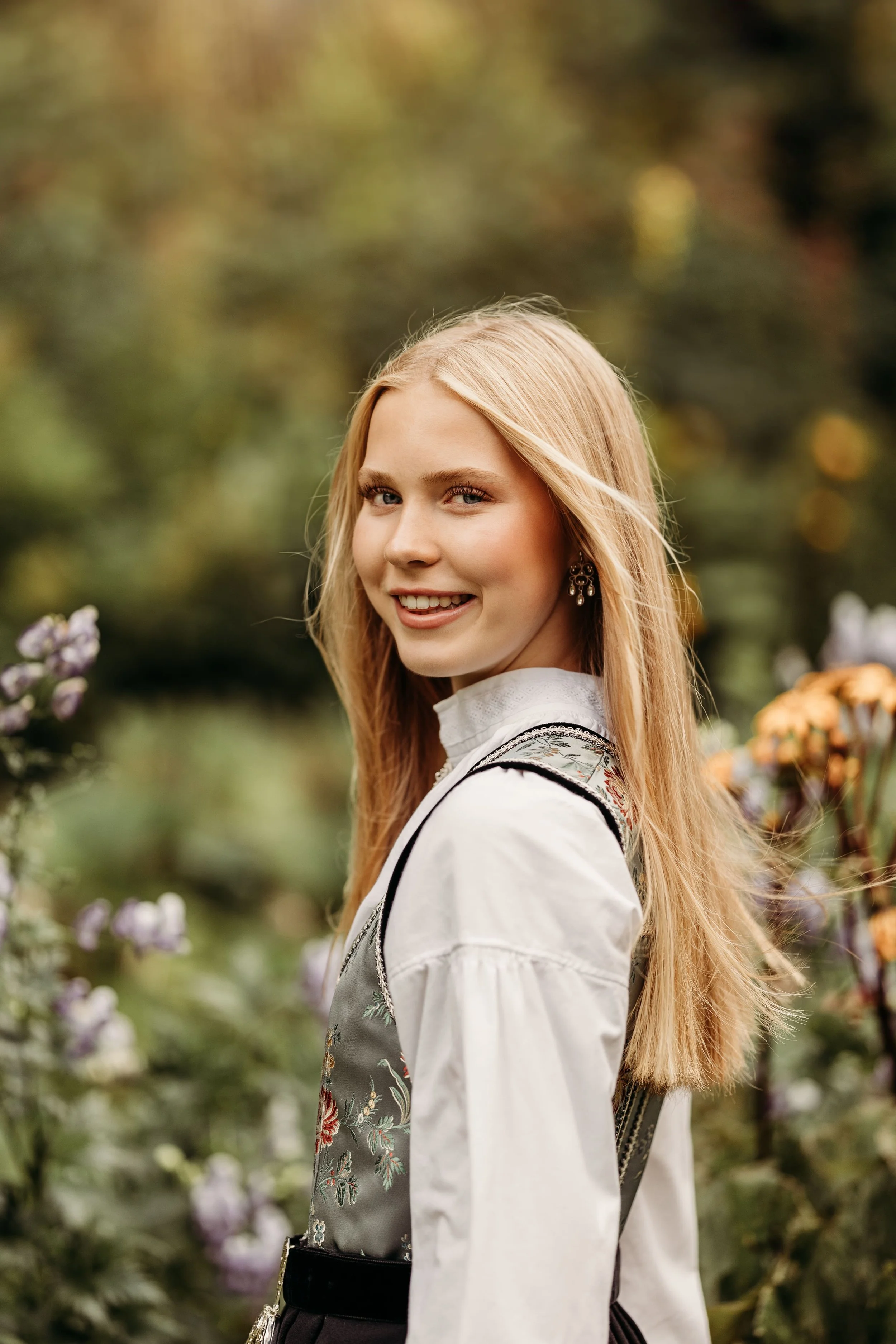 En smilende kvinne med langt blondt hår, iført tradisjonell folkedrakt, står i en blomsterhage med blomster og blader i bakgrunnen.