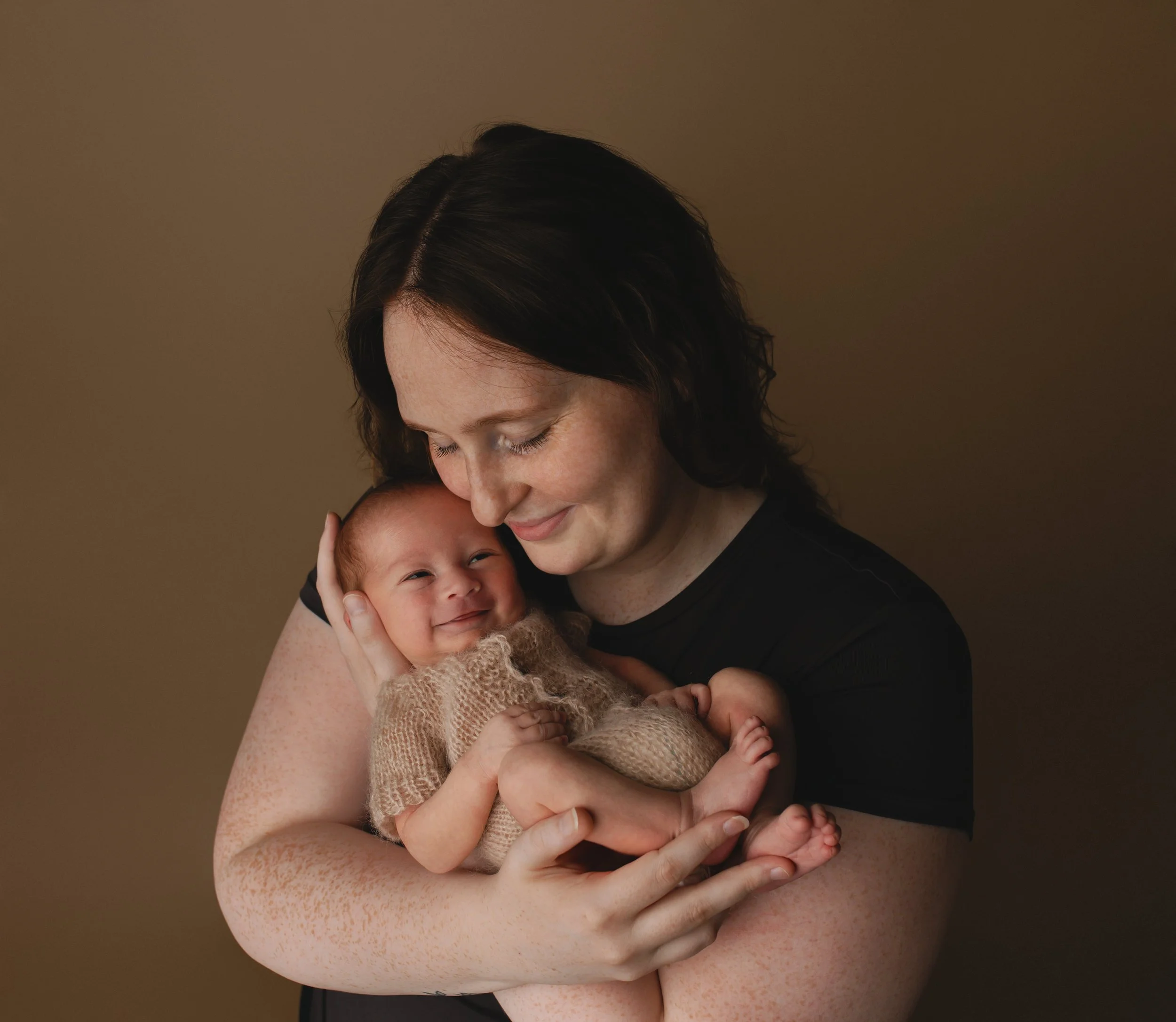 En kvinne med mørkt, krøllete hår holder en baby som smiler. Bakgrunnen er beige.