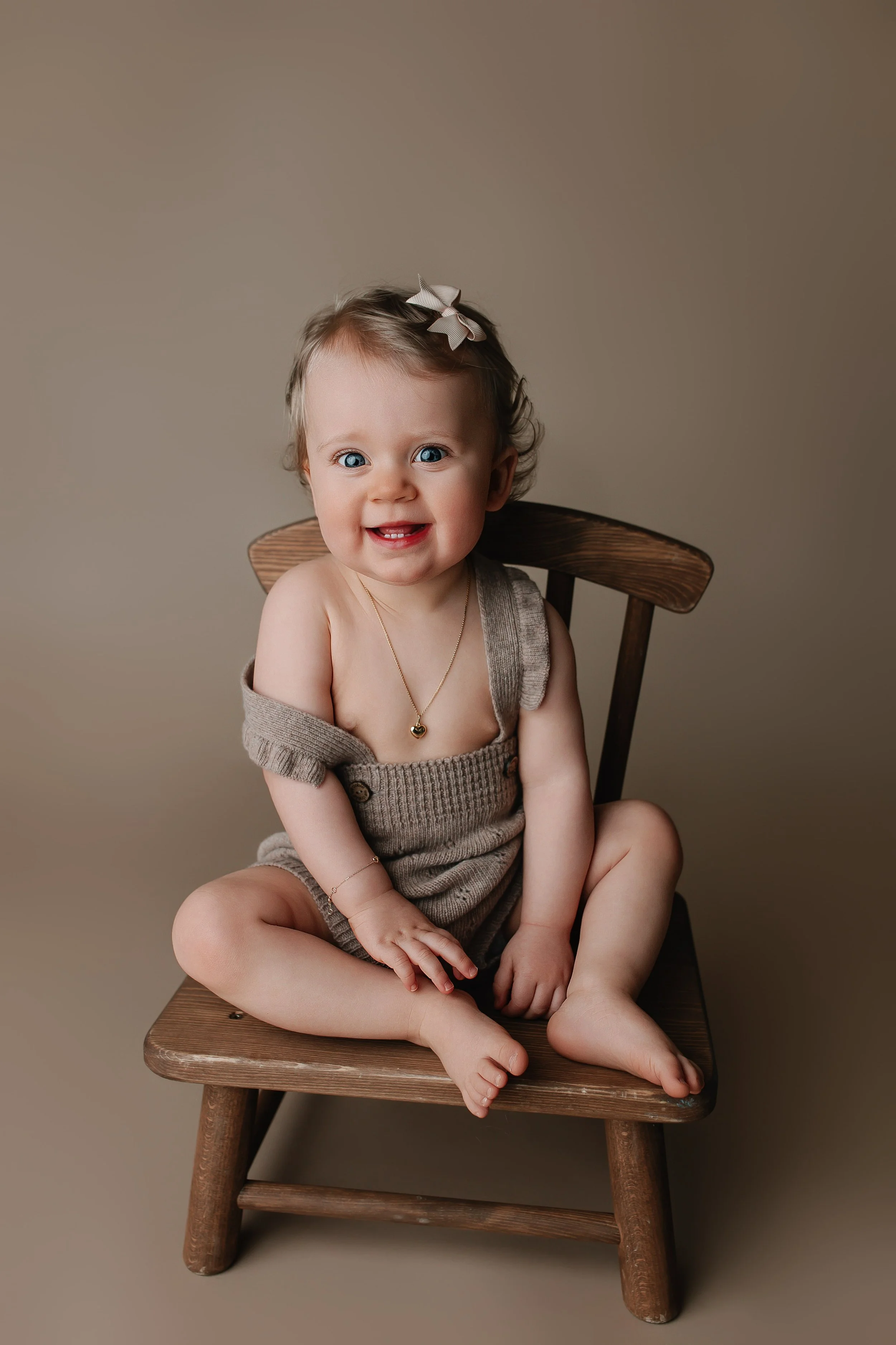 En smilende babyjente med blå øyne, kort brunt hår med en hvit hårbøyle, sitter på en liten trestol. Hun har på seg en beige ullkåpe med nesten ingen klær under, og har på seg smykker, inkludert en kjede med en hjertevedheng og et armbånd.