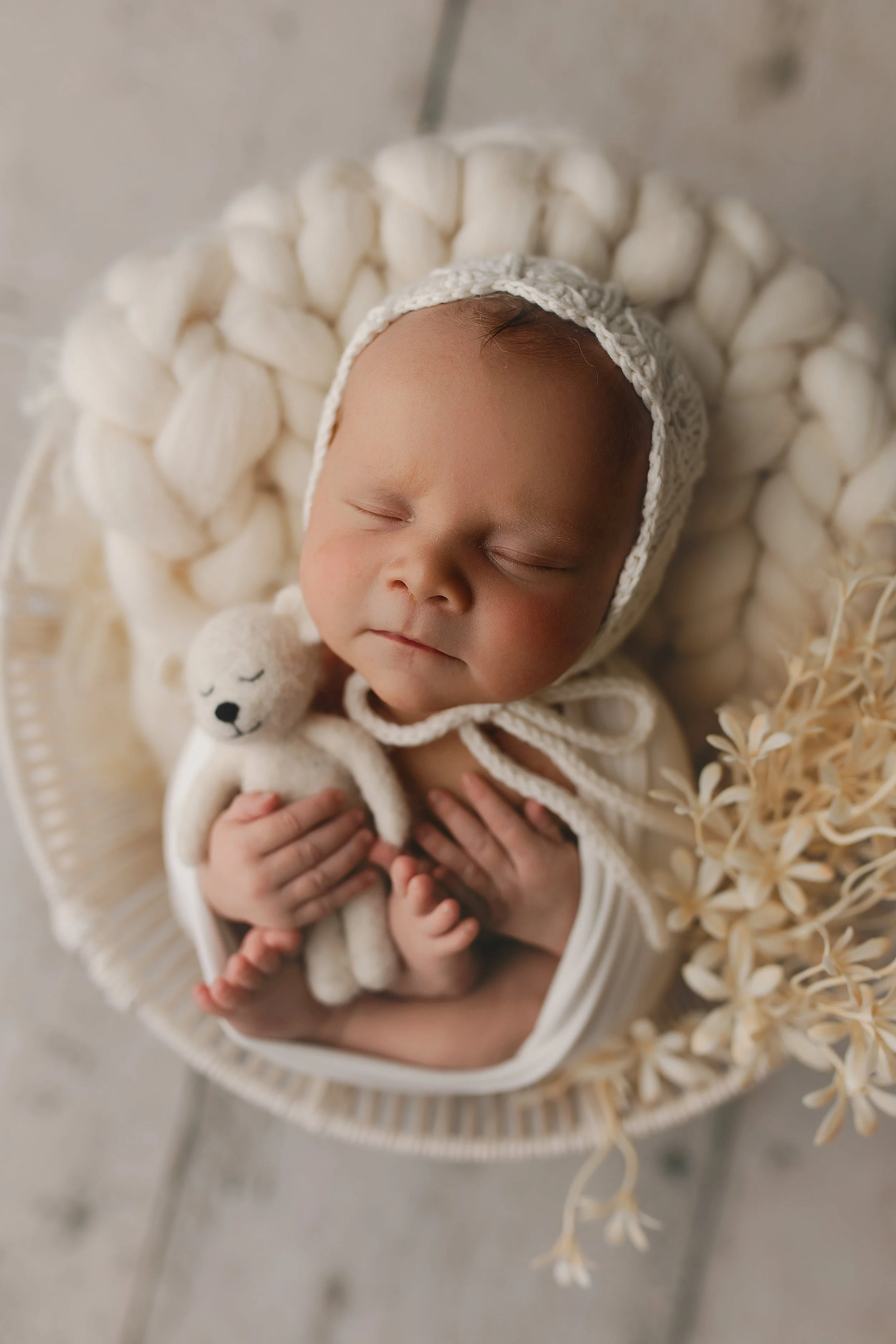En sovende baby med en hvit strikket lue, som holder en liten hvit bamse, liggende i en kurv med hvite vattpinner og tørkede blomster rundt.