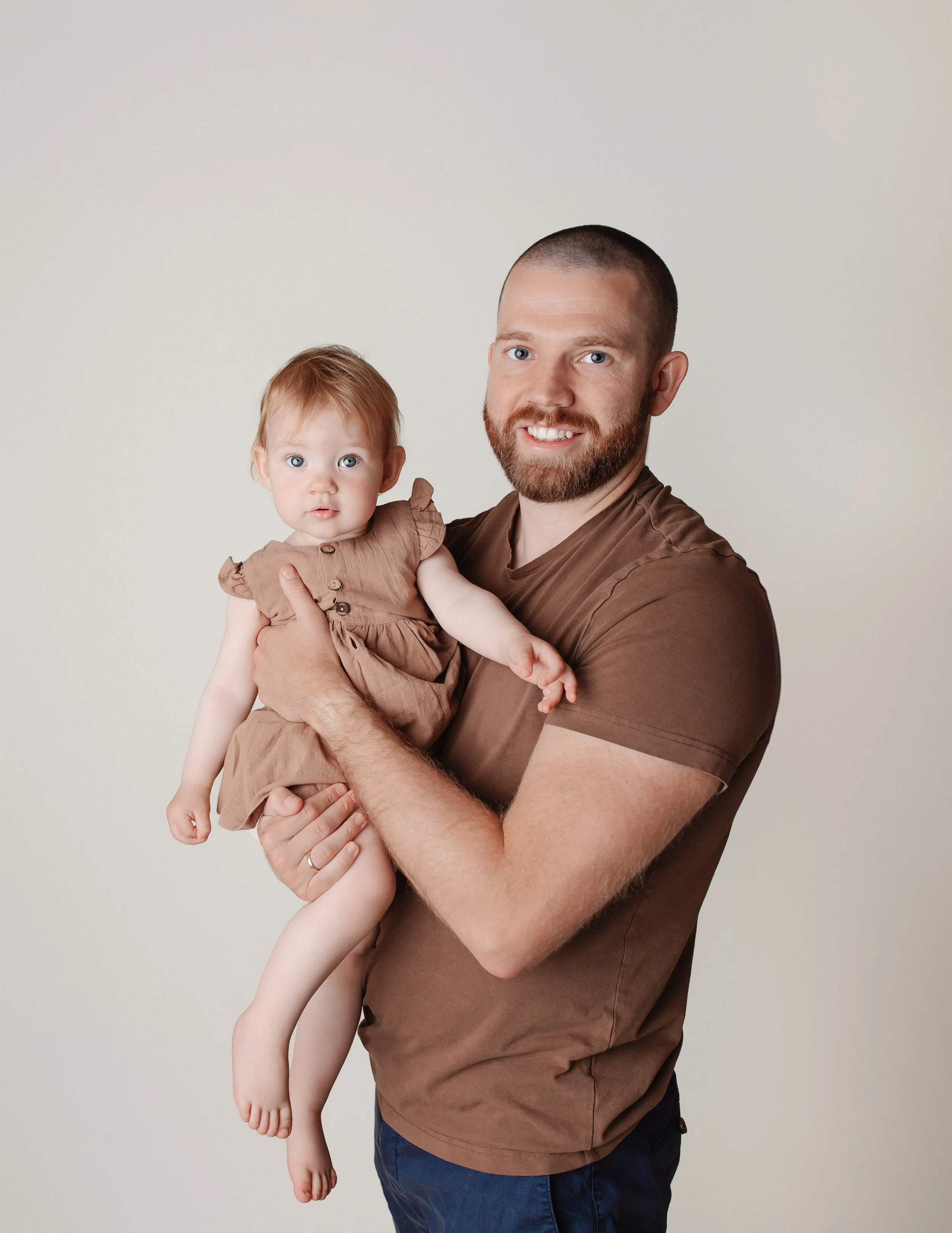 En mann med bart som smiler, holder en liten jente med rødt hår og blå øyne.