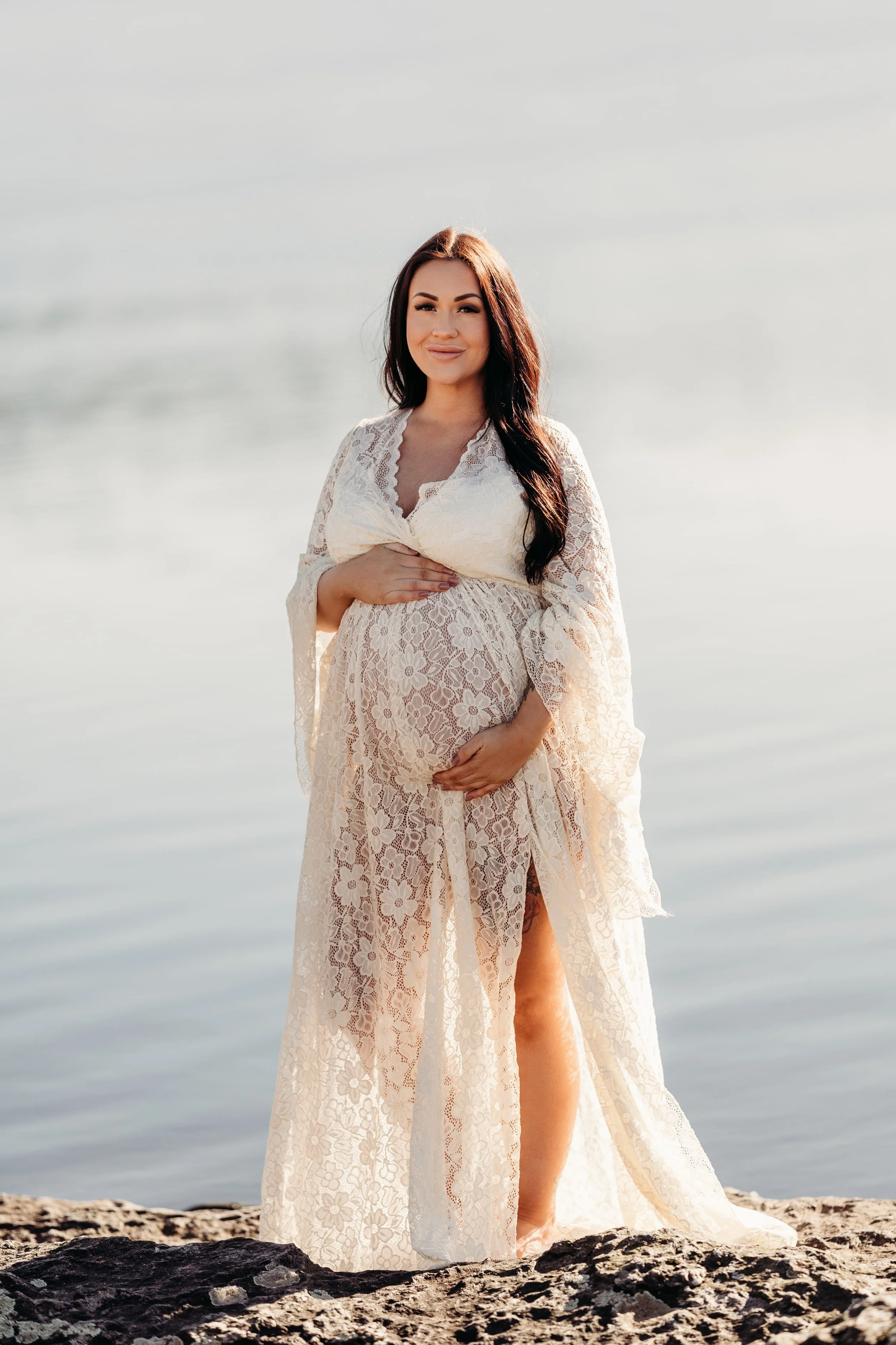 Gravid kvinne i hvit blondeklær står på en stein ved vannet.