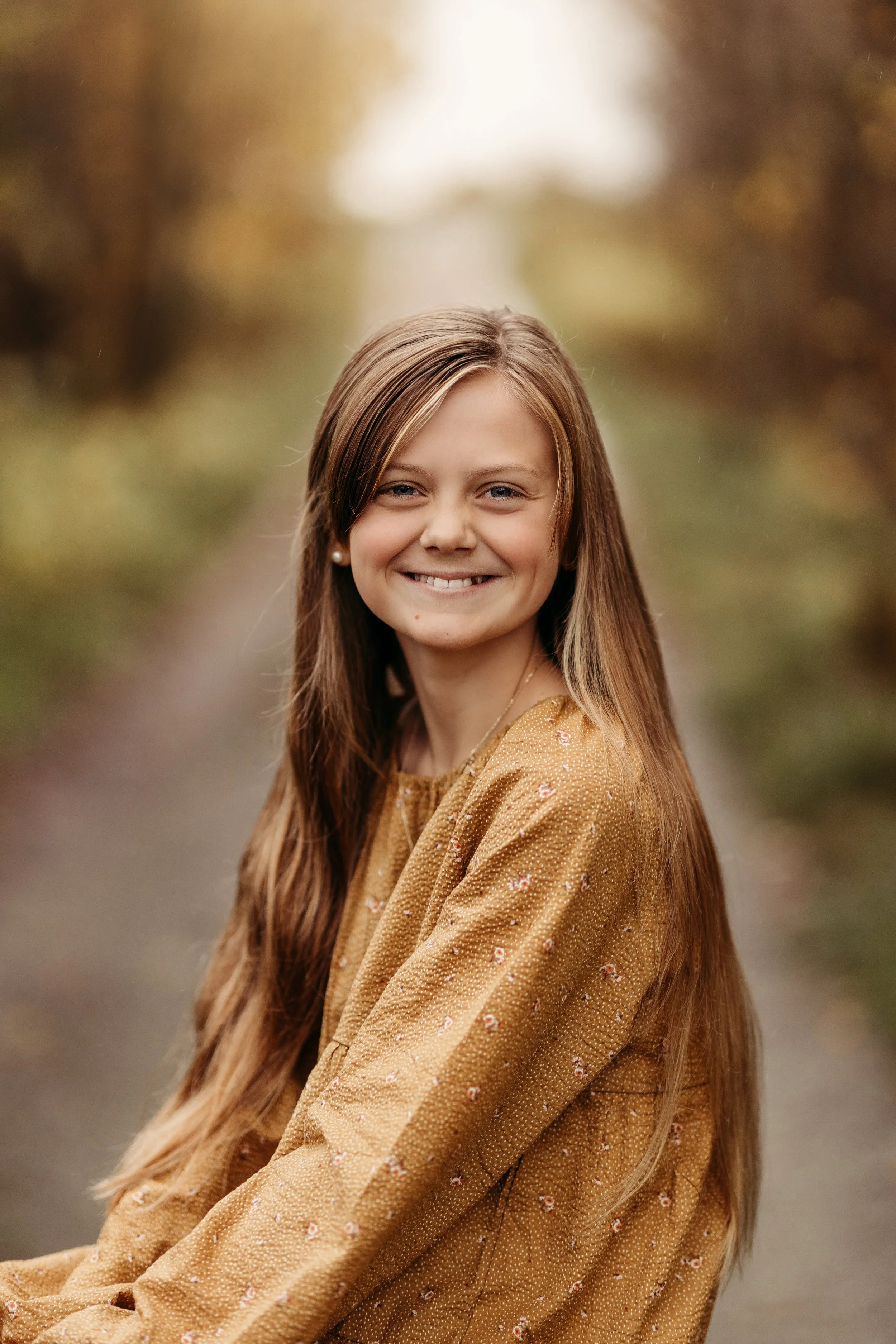 En smilende ung kvinne med langt brunt hår, sittende utendørs på en gangsti med høstlignende bakgrunn.