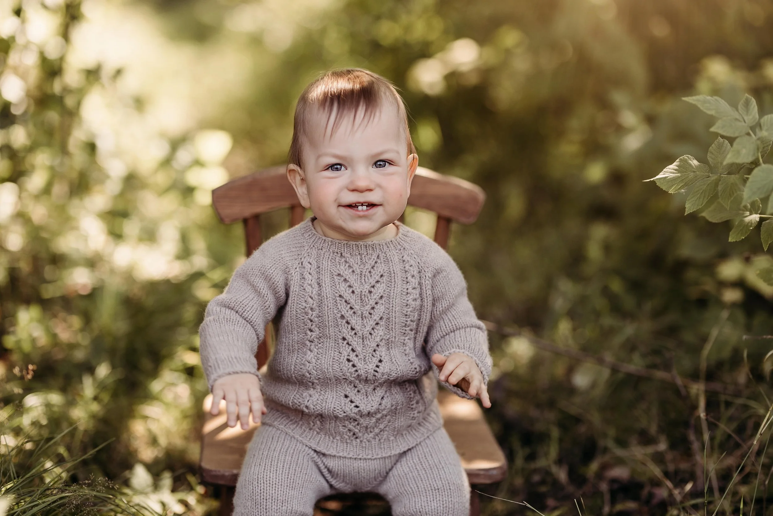 En liten baby i lys beige strikkeplagg som sitter på en trestol i en skogsmiljø om høsten.