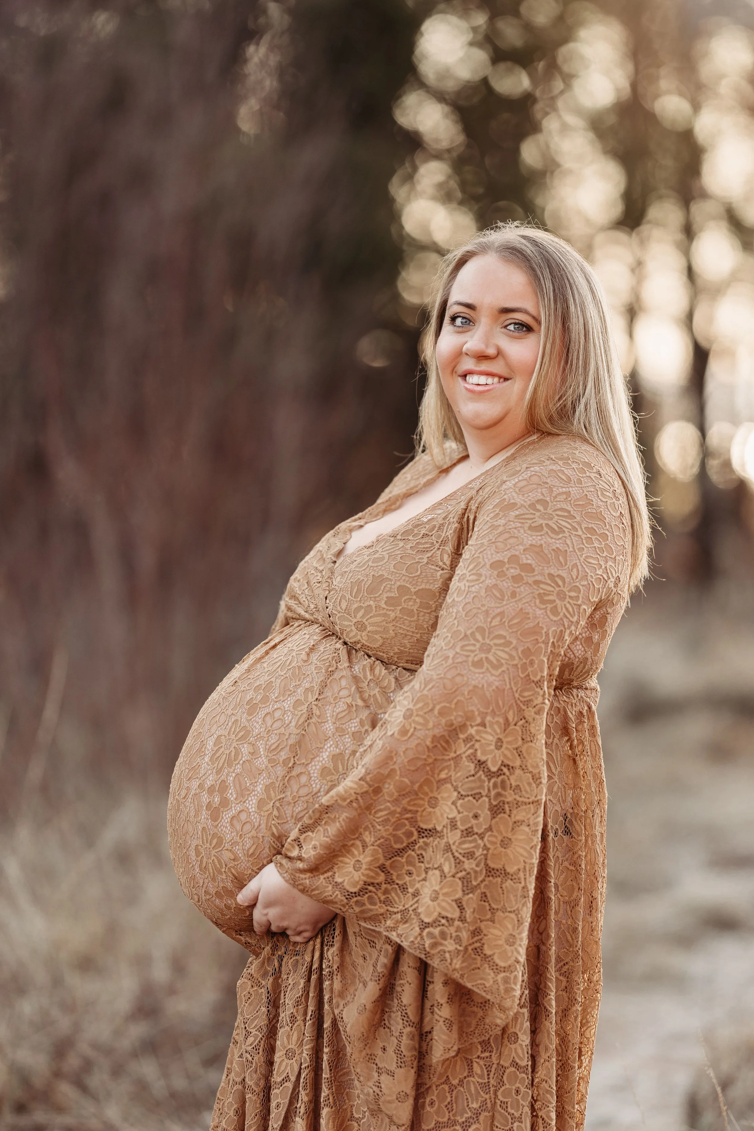 En gravid kvinne i en beige, blomstret kjole står utendørs med bladverk i bakgrunnen, smilende mot kamera.