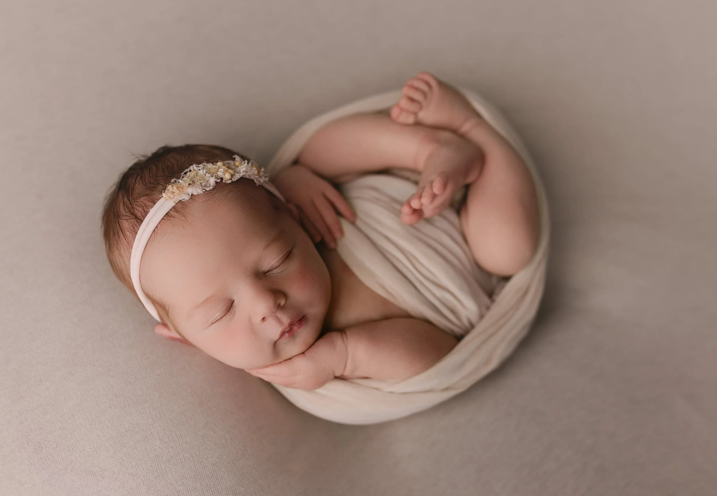 Søvnende baby med blomsterhodebånd, pakket inn i et lysebeige stoff, liggende på en myk overflate.