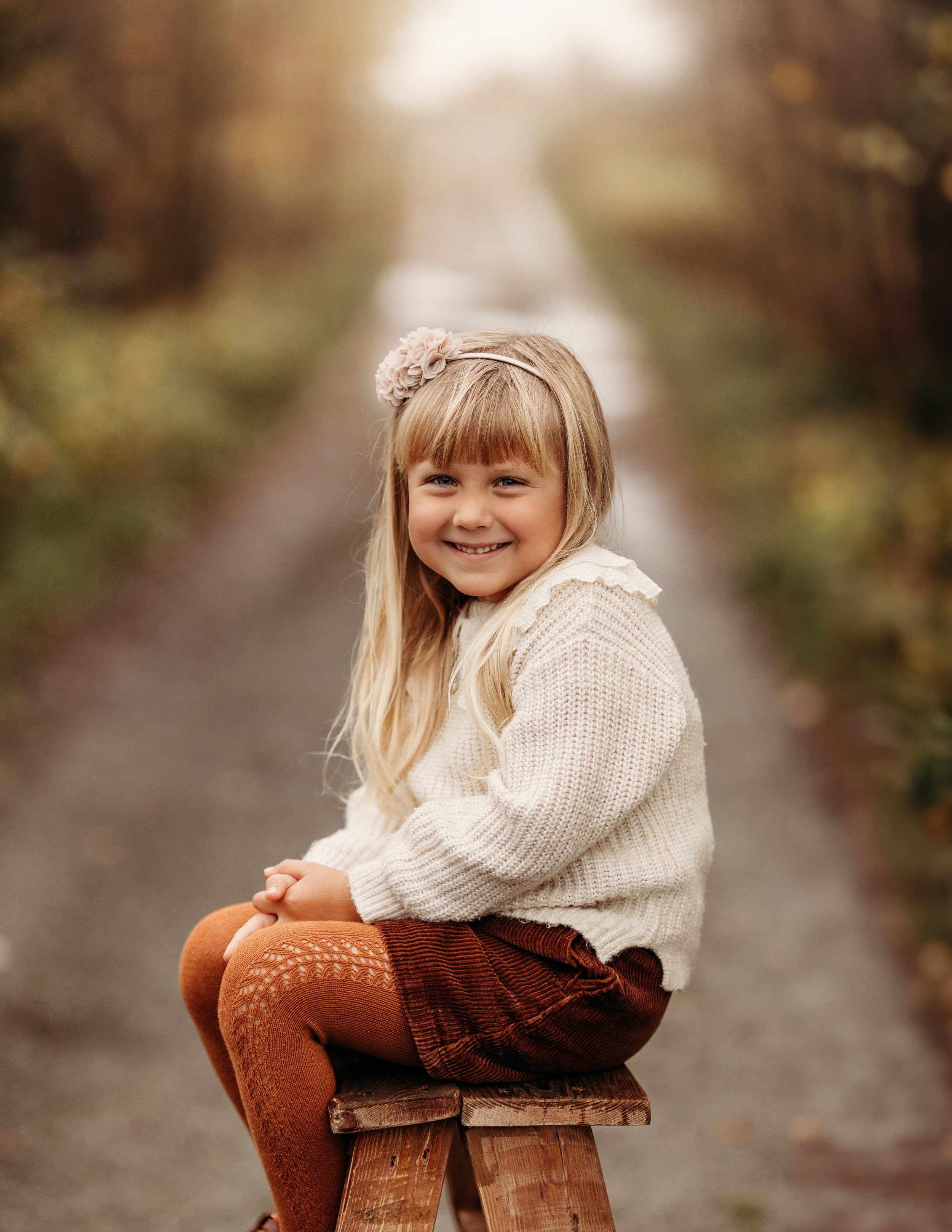 En liten jente sitter på en trebenk på en sti, smilende mot kamera, omgitt av høstblader.