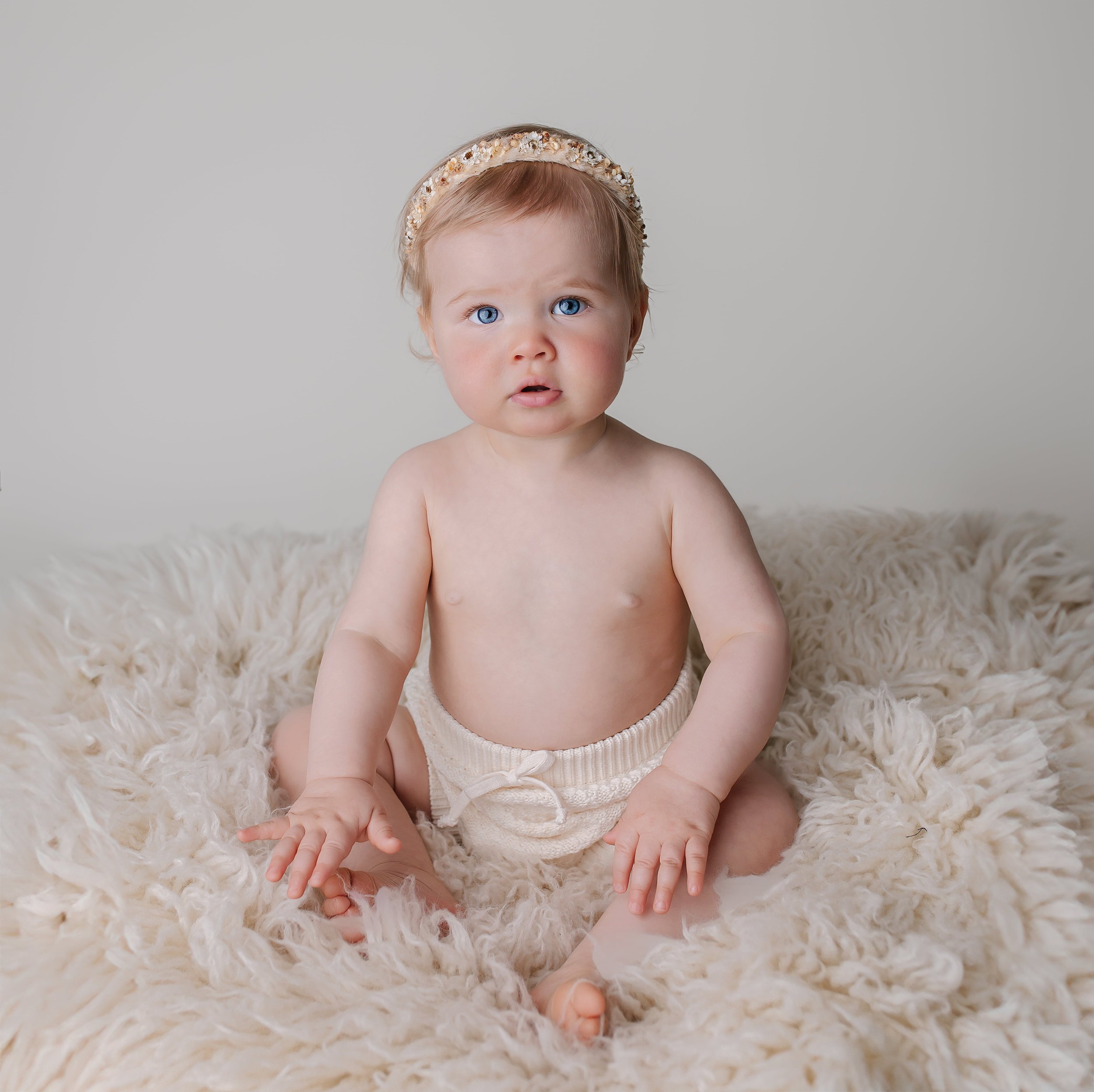 En liten baby med blå øyne som sitter på en myk, hvit pelsdekt overflate.