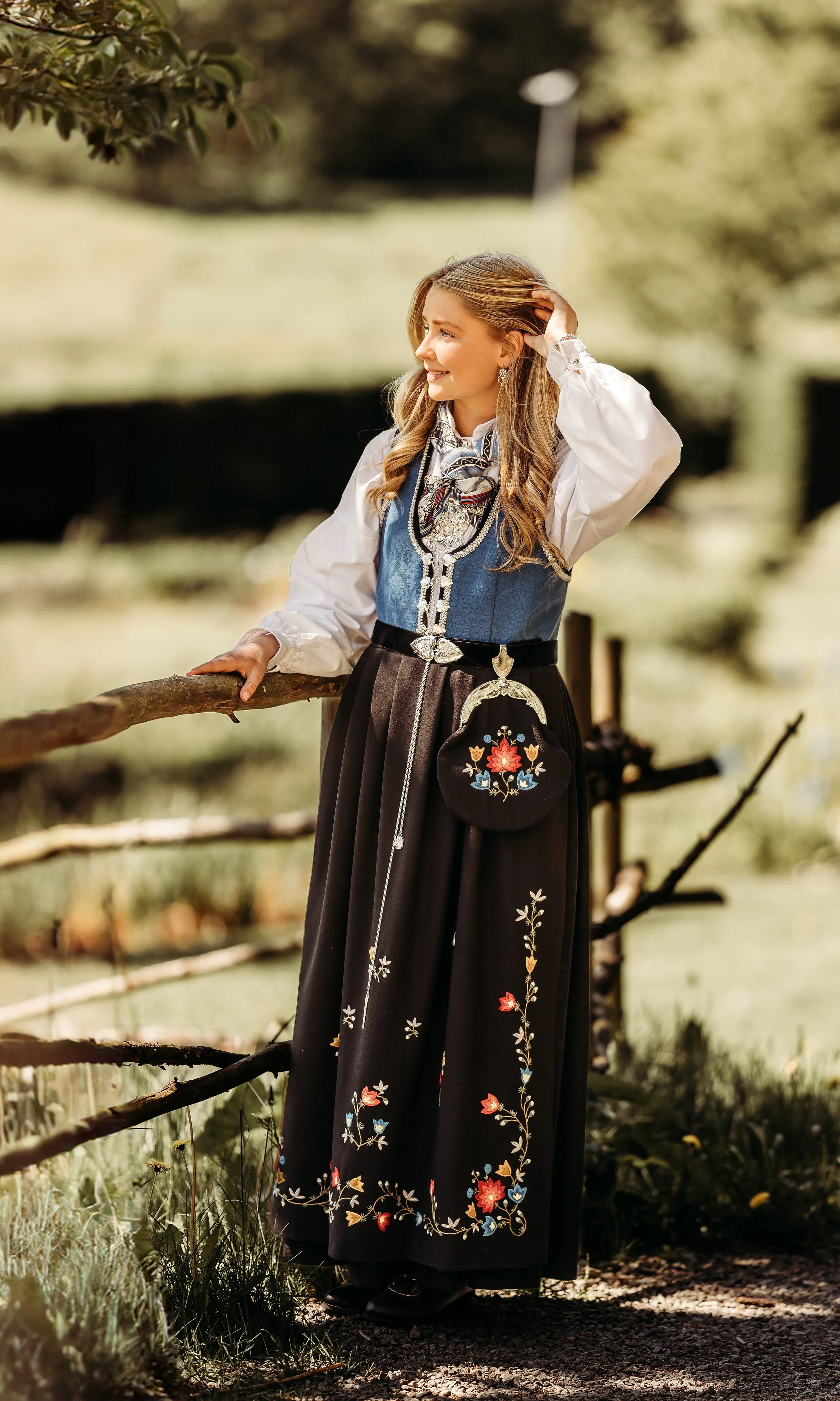 Kvinne kledd i tradisjonell norsk bunad, står ved en hagegjerde med blader og trær i bakgrunnen.