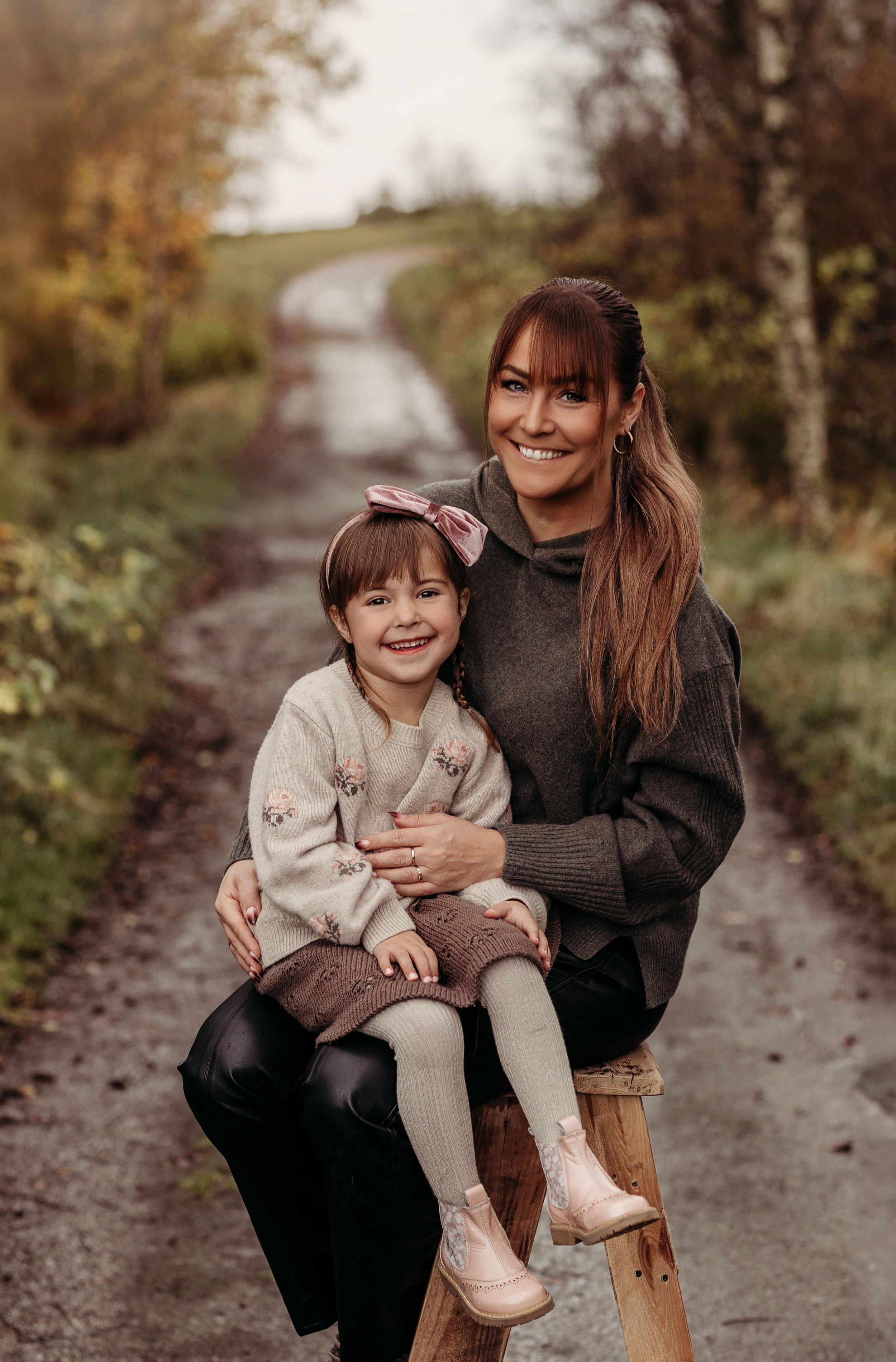 En kvinne og et barn som smiler, sittende på en trebøtte i en natursti omgitt av høstfarger.