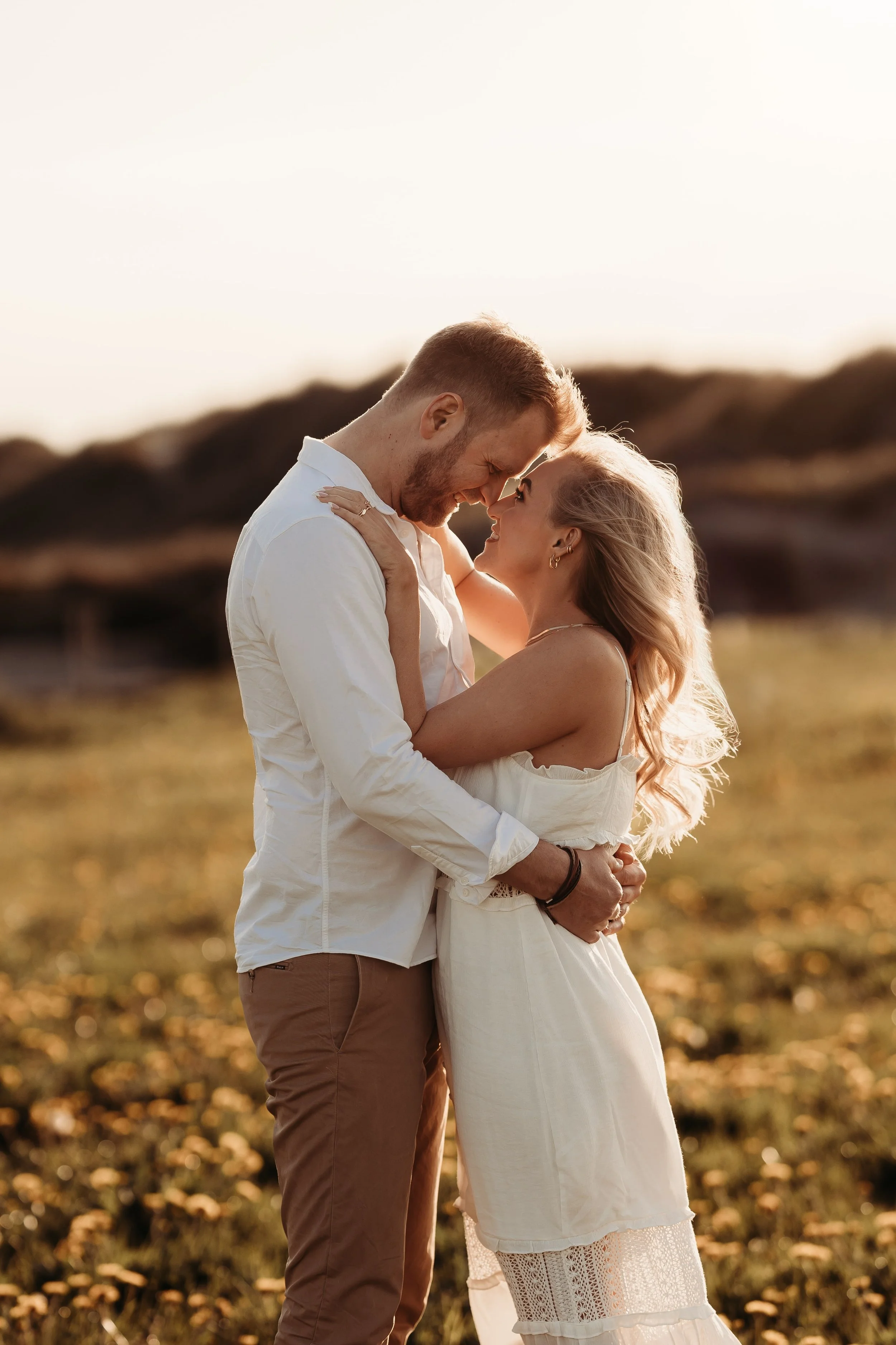 Et kjærlighetspar står tett sammen, smiler og ser inn i hverandres øyne i utendørs landskap under solnedgang.