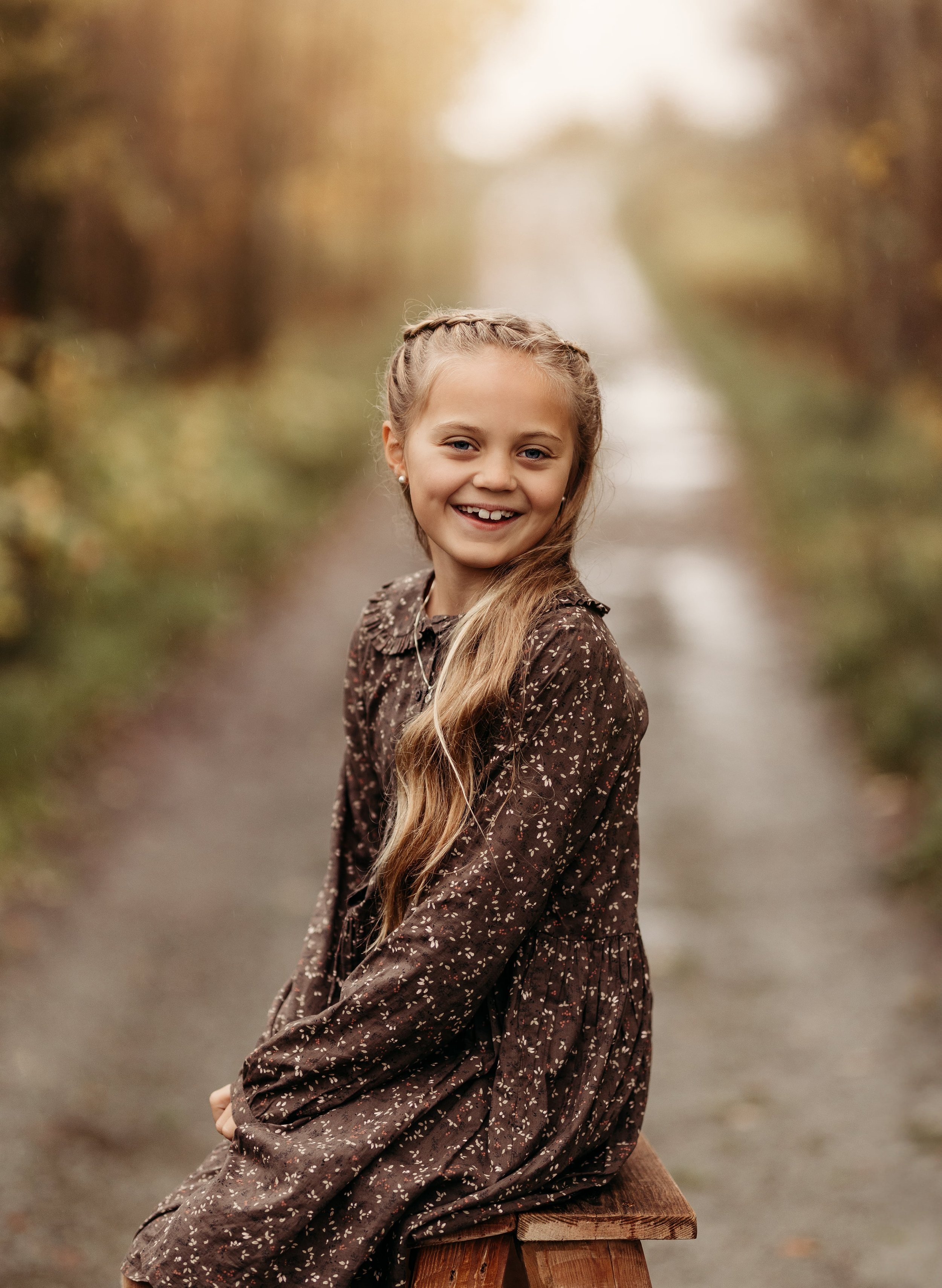 Ung jente med langt blondt hår som smiler, sittende på en trebord i en skogssti med høstfargede trær i bakgrunnen.