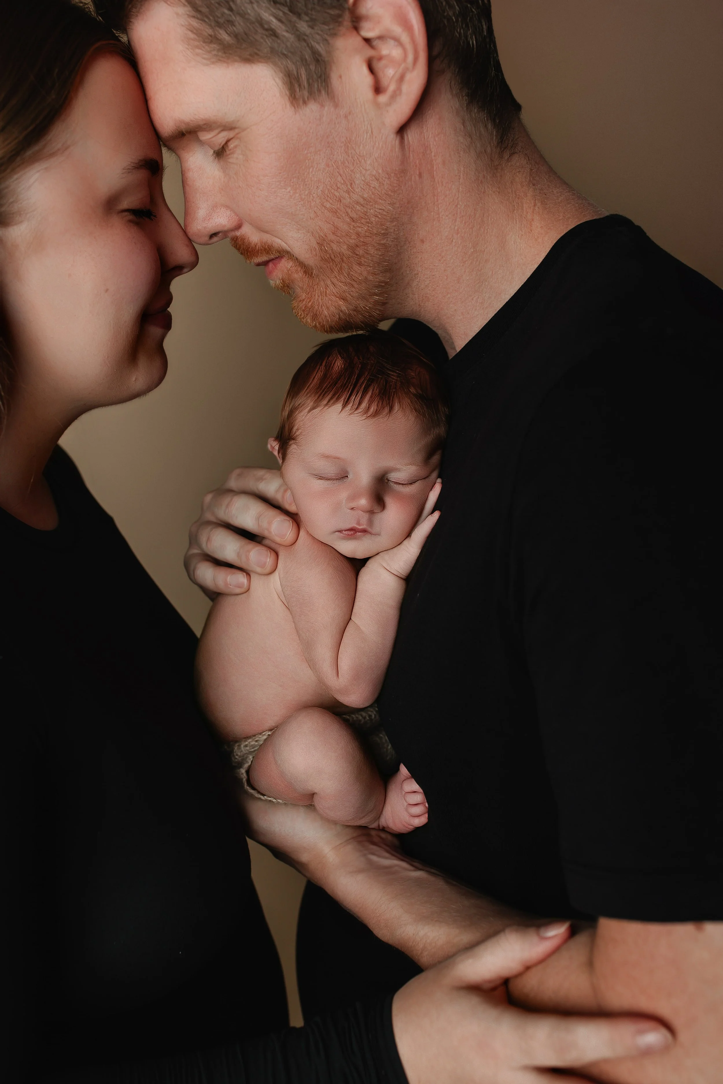 Et nyfødt barn sovende i armene på faren, mens mor og far har panna mot panna i en kjærlig og intim øyeblikk.