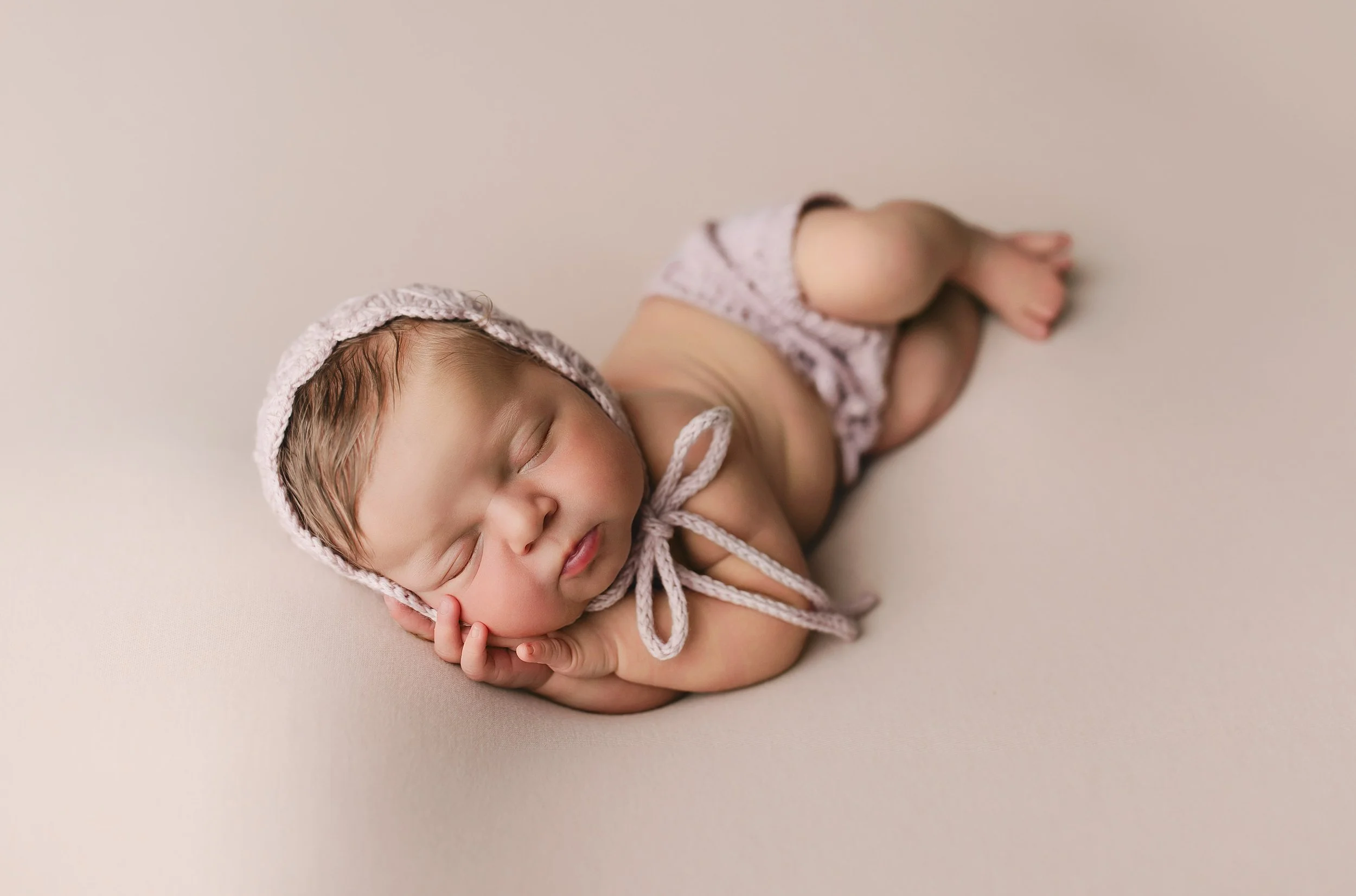 Nydelig nyfødt baby sovende med hodet støttet av hånden, iført en rosa strikket hette og truse, liggende på en myk, lys bakgrunn.