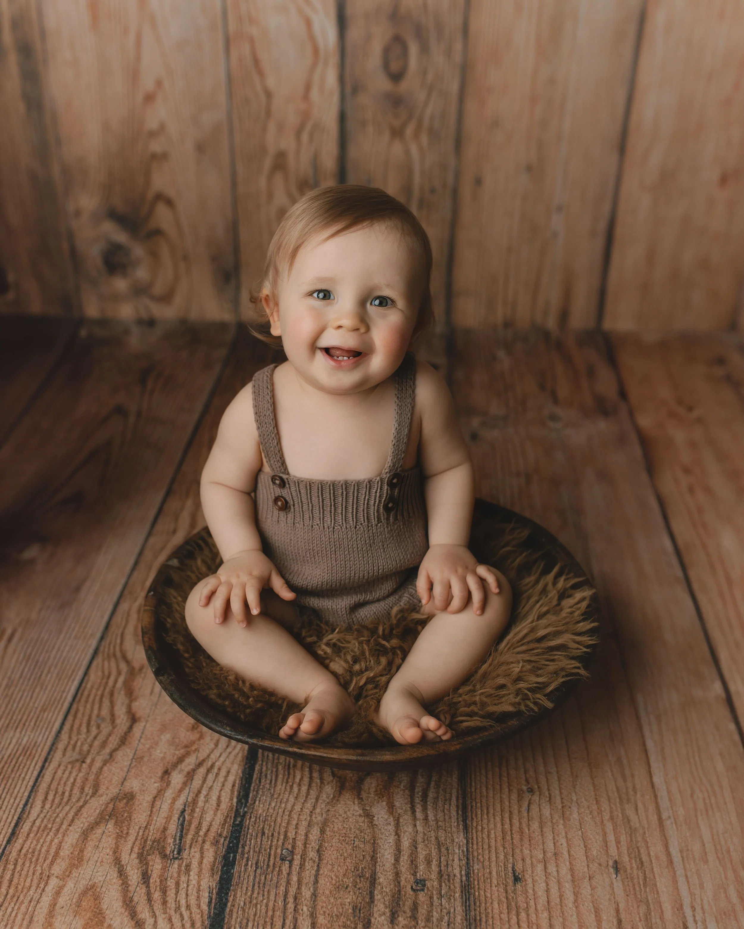 Liten gutt sitter i en rund skål med pels på en tregulv med trepanel bakgrunn.